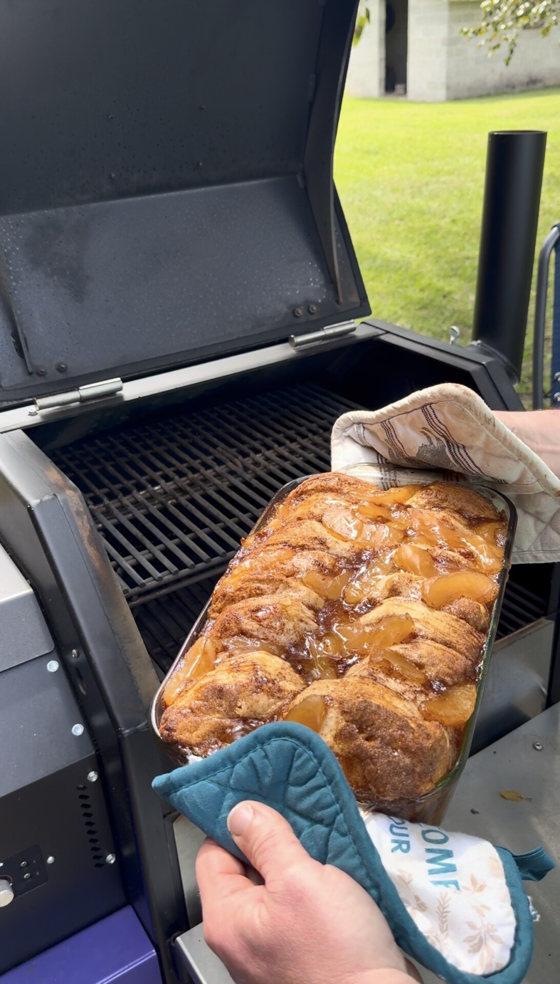 A person uses oven mitts to hold a freshly baked peach cobbler in a glass dish, taking it off a grill outdoors. The cobbler is golden brown with visible peach slices and a caramelized topping.