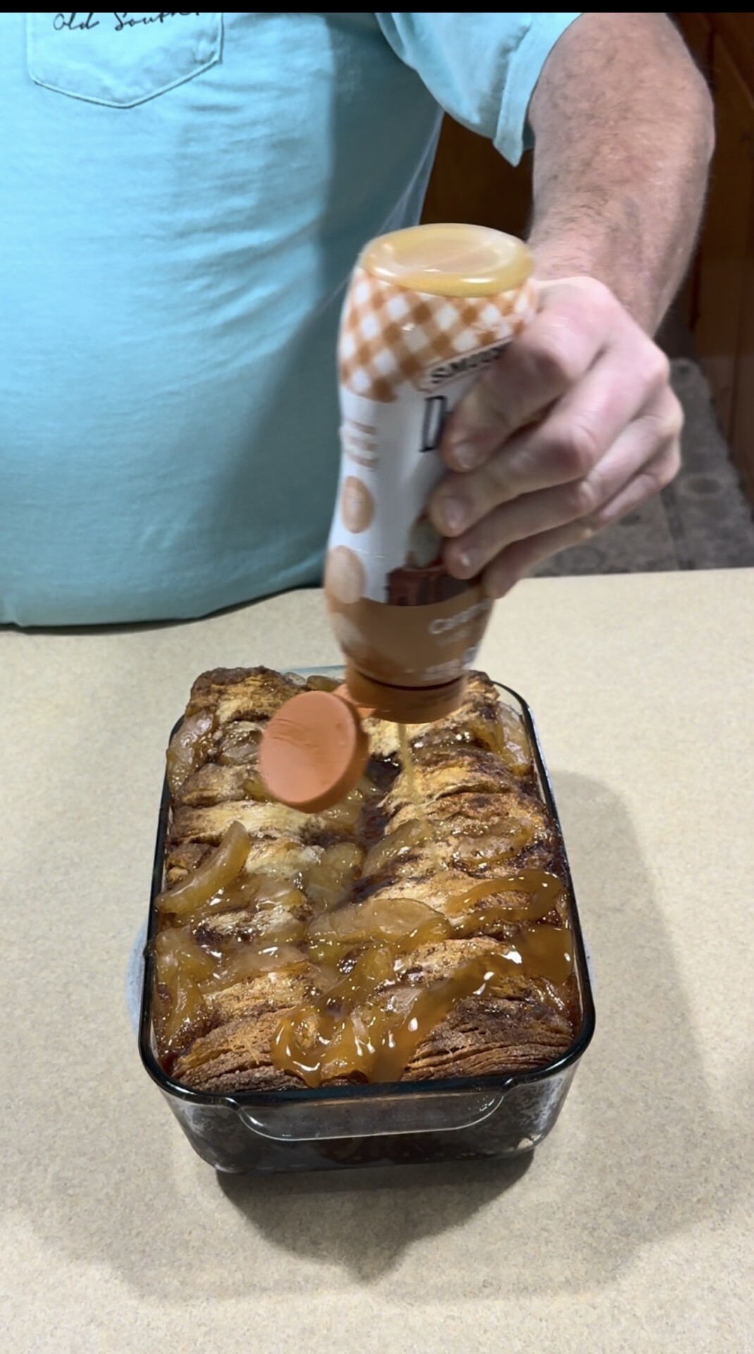 A person in a light blue shirt pours caramel sauce over a baked dessert in a glass dish, which appears to be a pull-apart bread or pastry, sitting on a countertop.