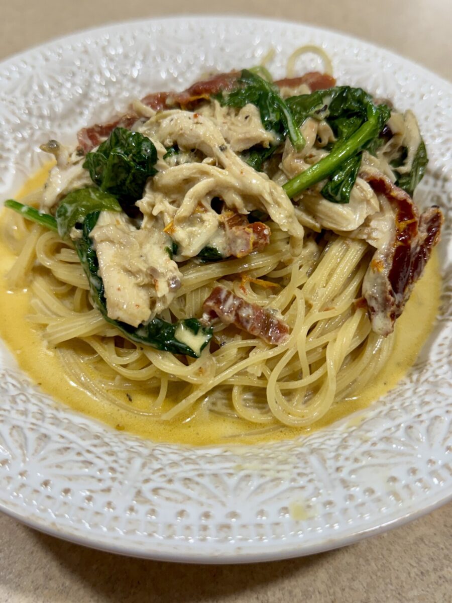 A plate of pasta topped with shredded chicken, spinach, sun-dried tomatoes, and a creamy sauce, served on a decorative white dish.