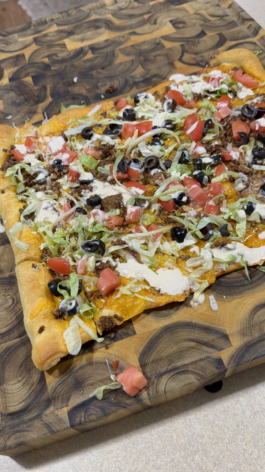 A rectangular taco-style pizza topped with ground meat, shredded lettuce, diced tomatoes, sliced black olives, and a drizzle of white sauce on a wooden cutting board.