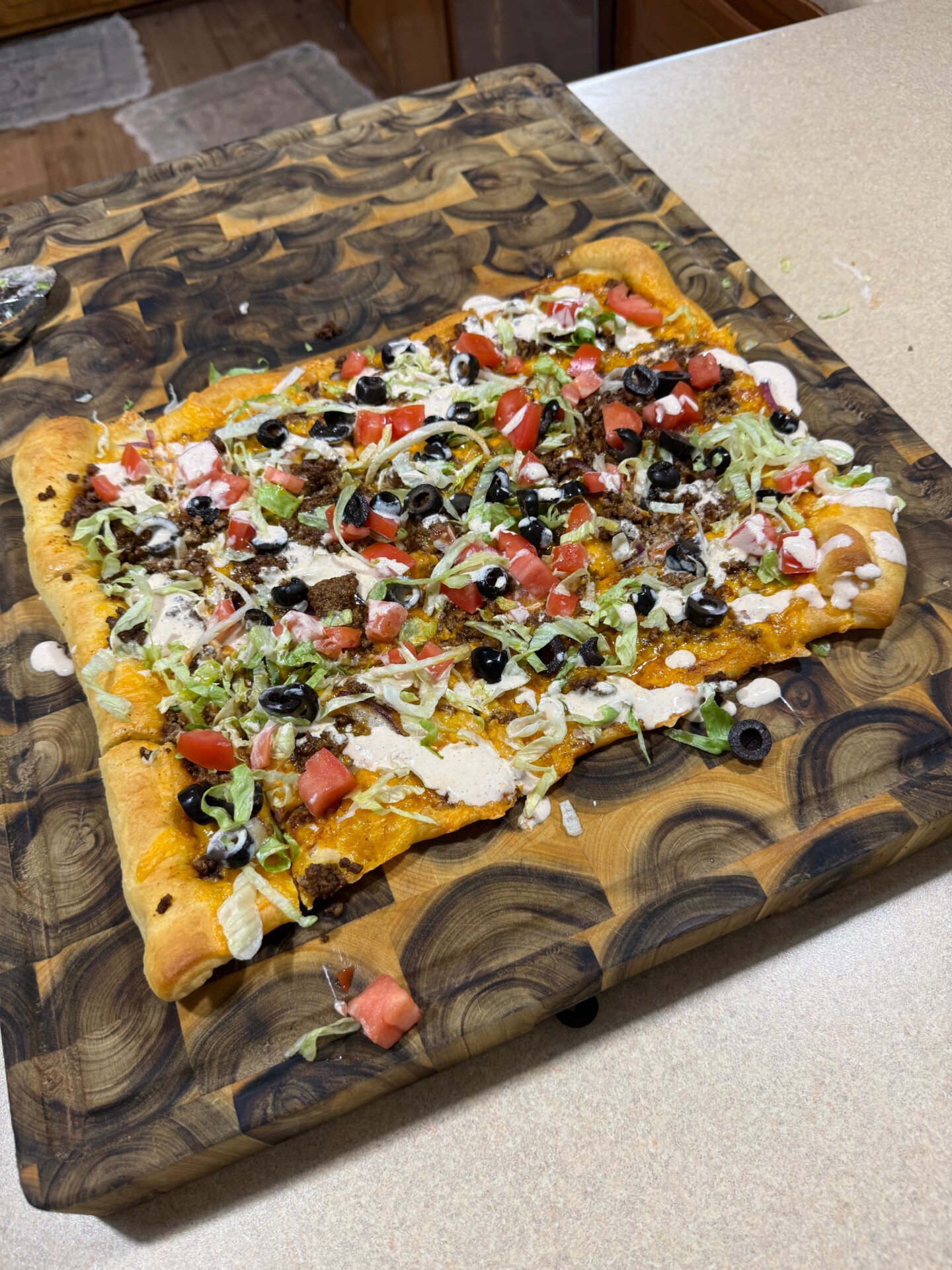 Rectangular taco pizza topped with ground meat, shredded lettuce, diced tomatoes, sliced black olives, and a drizzle of creamy sauce on a wooden cutting board.