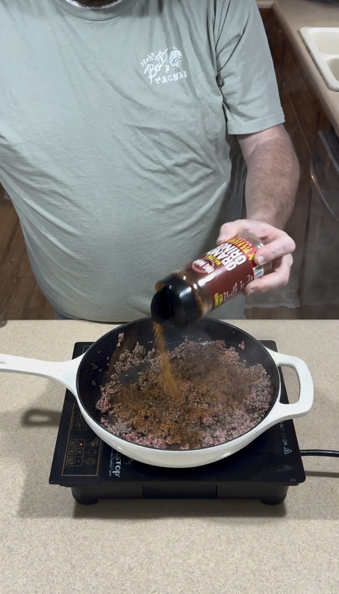 A person in a light green t-shirt pours seasoning from a bottle into a skillet containing browning ground meat on a portable induction burner.