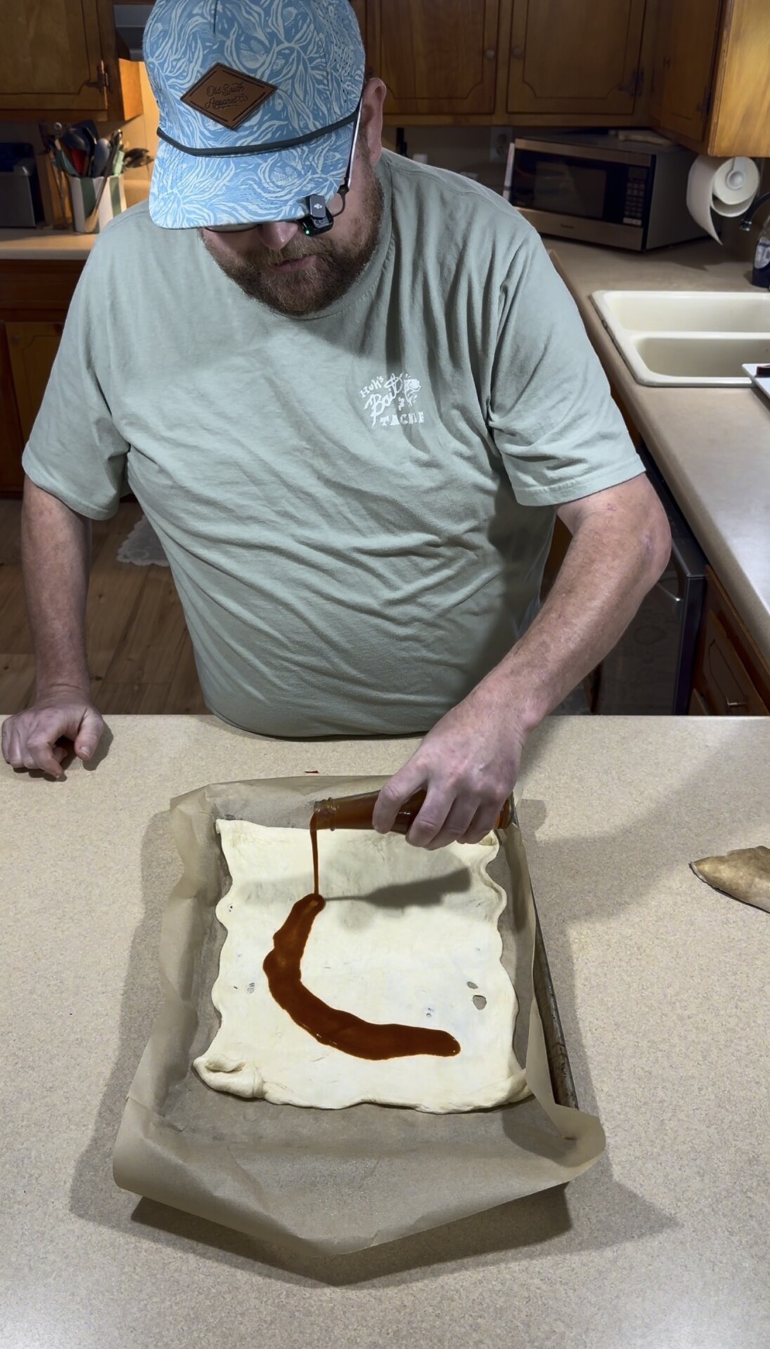A man in a baseball cap pouring barbecue sauce onto a rectangular sheet of raw dough on a baking pan.