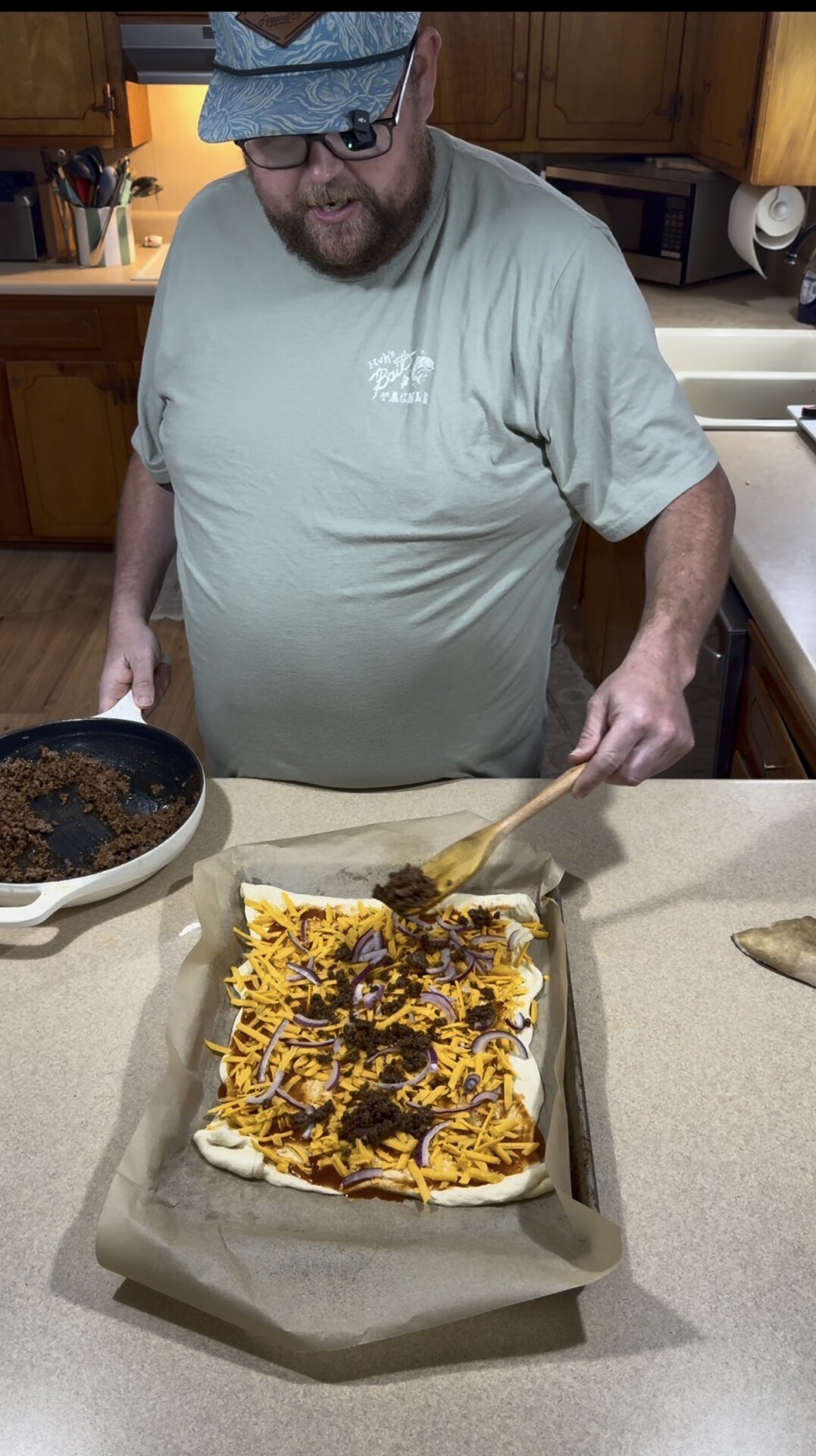 A person using a wooden spoon to spread cooked ground meat onto a rectangular pastry base topped with shredded cheese and sliced red onions.