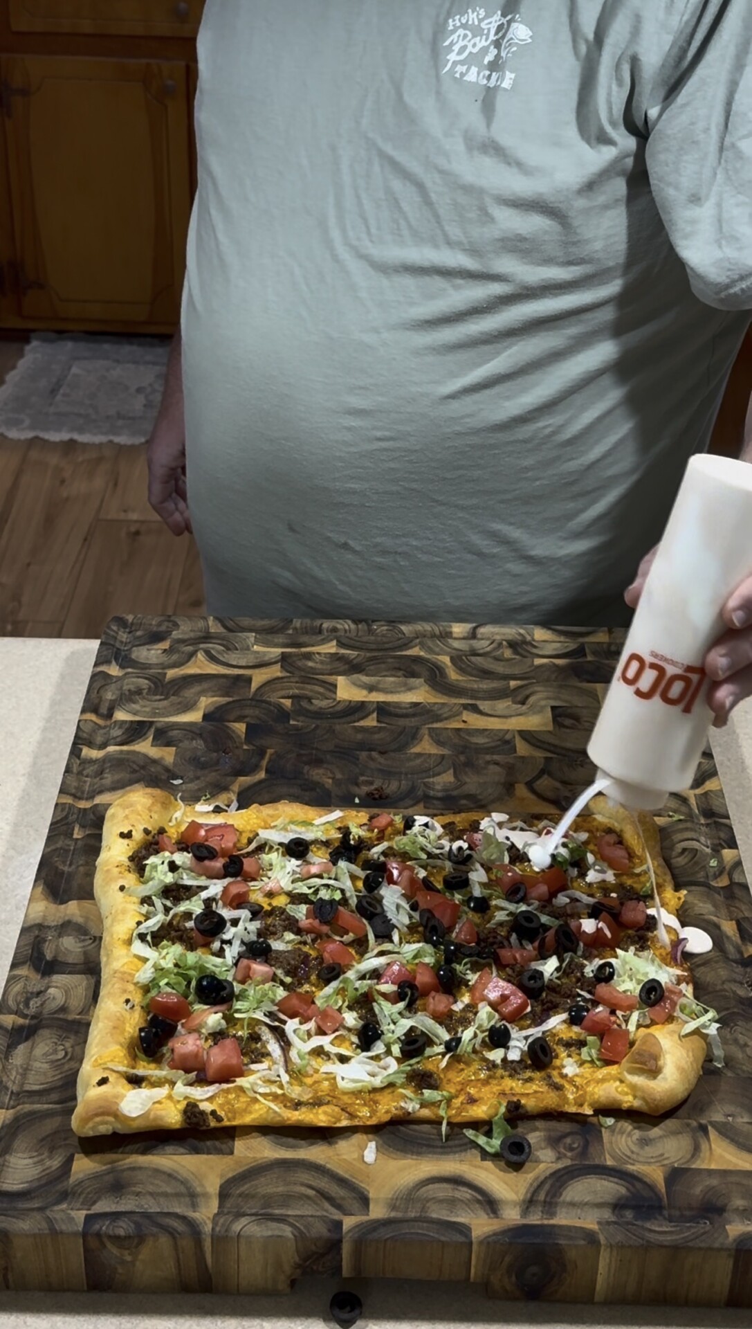 A person squeezing a white sauce onto a rectangular pizza topped with shredded lettuce, diced tomatoes, and black olives.