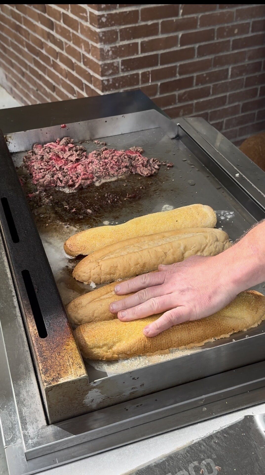 A hand presses three long bread rolls onto a flat-top grill next to a pile of cooking ground meat.