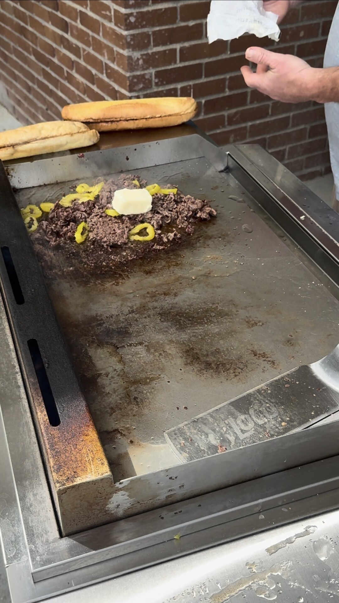A metal griddle cooking a pile of ground meat with sliced yellow peppers and a pat of butter, with two hoagie rolls resting on the side.