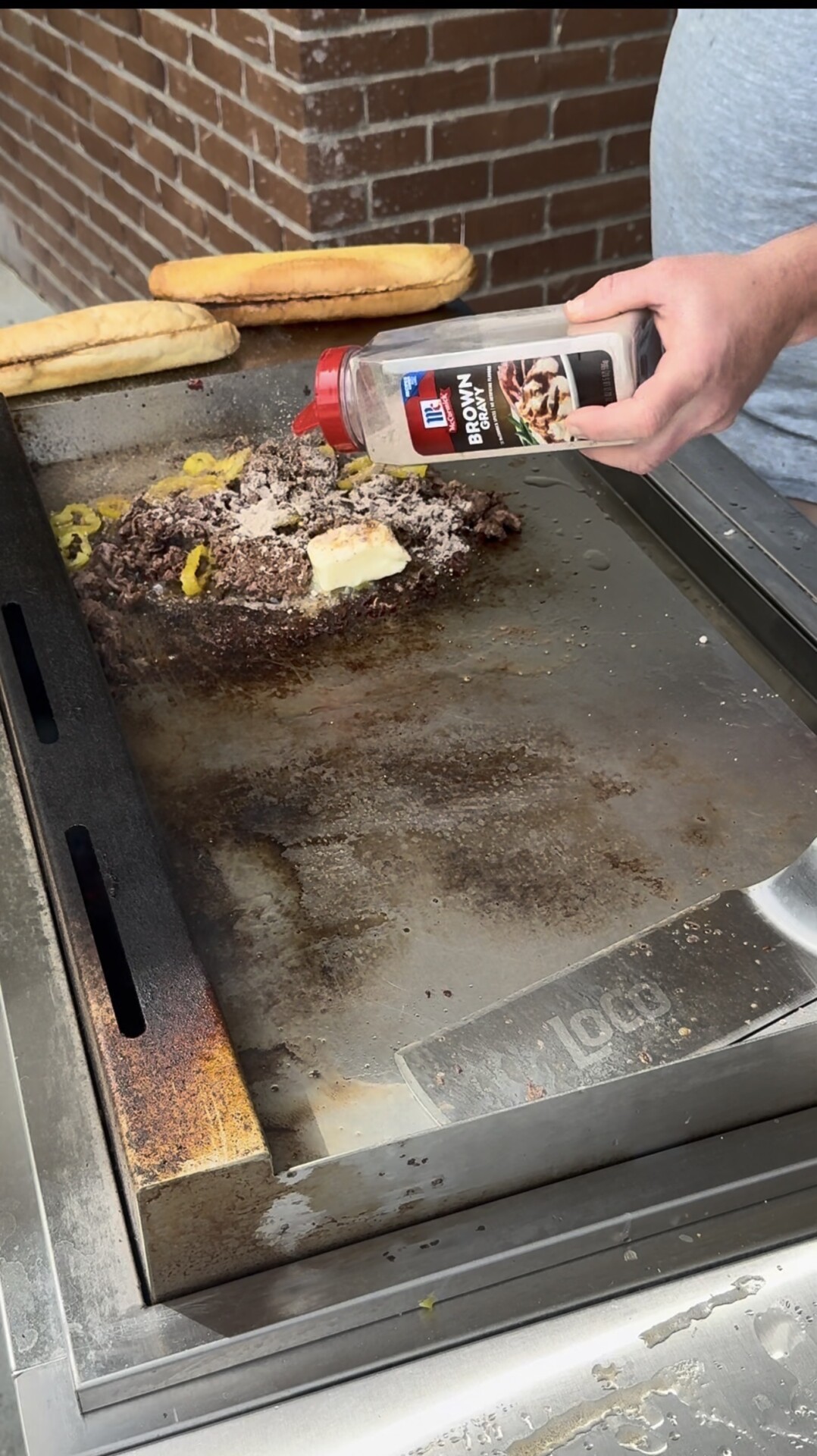 A hand sprinkles McCormick brown gravy mix onto a pile of chopped meat, peppers, and a pat of butter cooking on a flat-top grill, with two sandwich rolls sitting nearby.