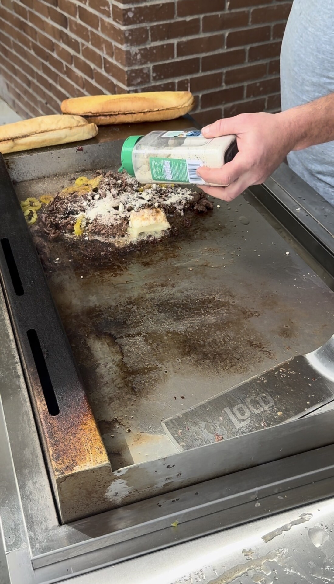 A person sprinkles seasoning from a shaker onto a pile of chopped meat, peppers, and cheese cooking on a flat-top grill.