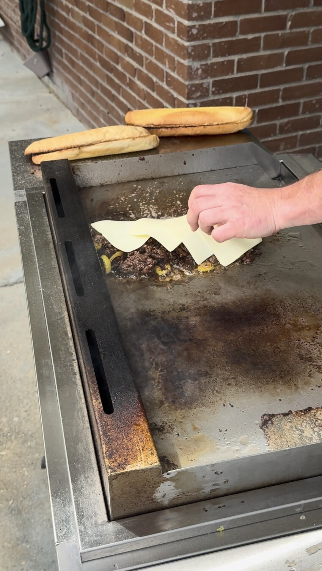 A hand places slices of cheese onto meat and vegetables cooking on a flat-top grill, with two toasted sandwich rolls resting nearby.
