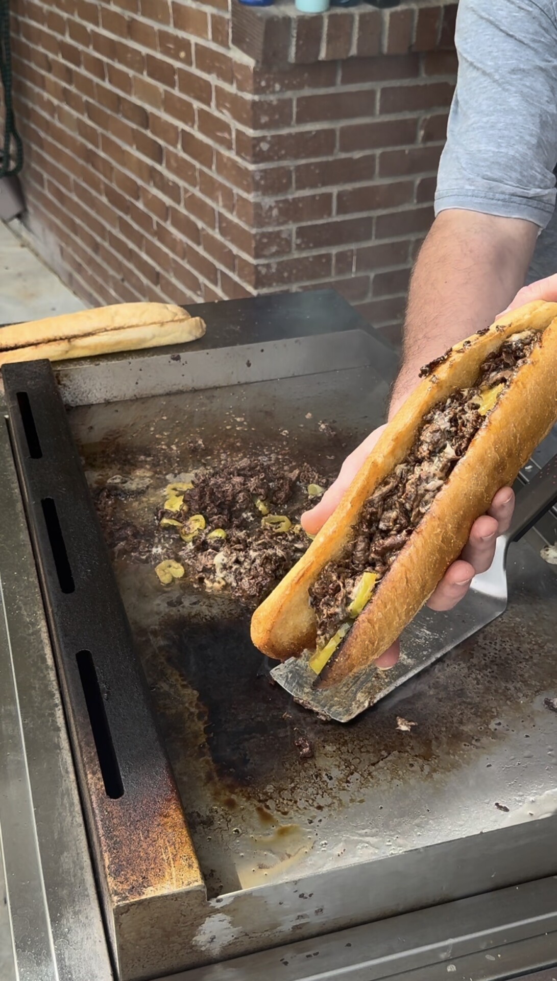 A hand holds a cheesesteak sandwich on a flat-top grill next to a pile of cooked meat and peppers.