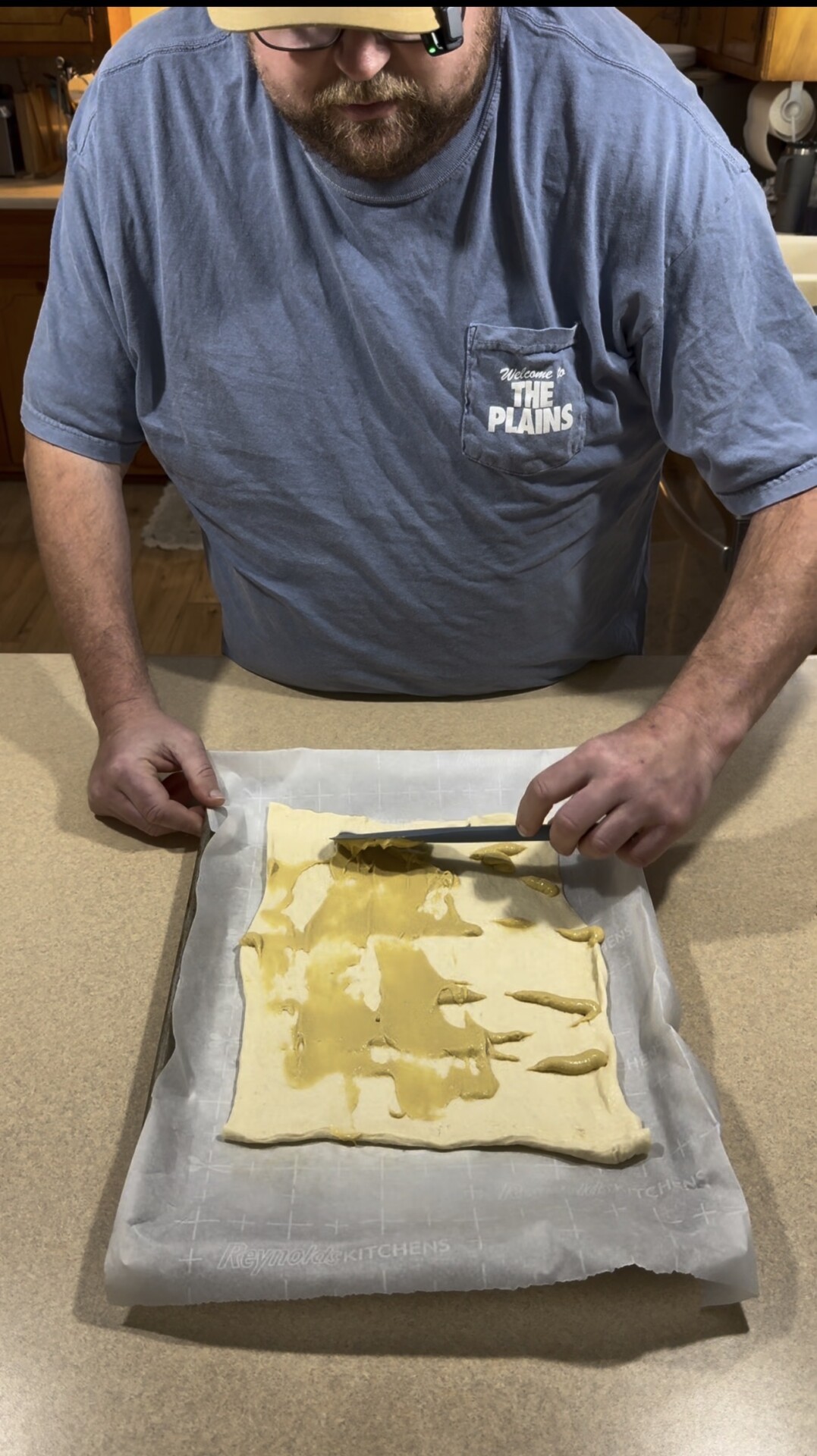 A person spreads mustard onto a sheet of puff pastry dough on a parchment-lined baking sheet.