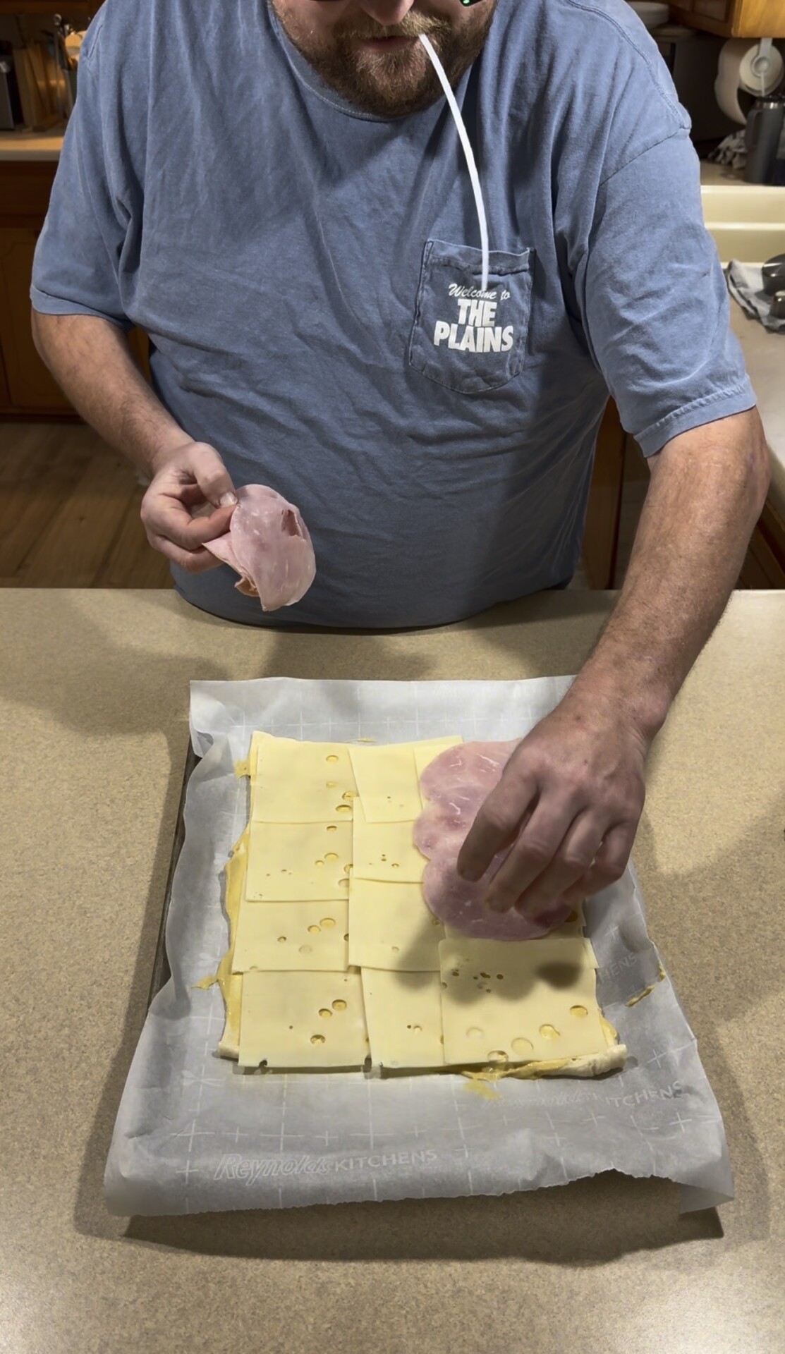 A person placing slices of ham onto a layer of Swiss cheese arranged on a parchment-lined baking sheet.