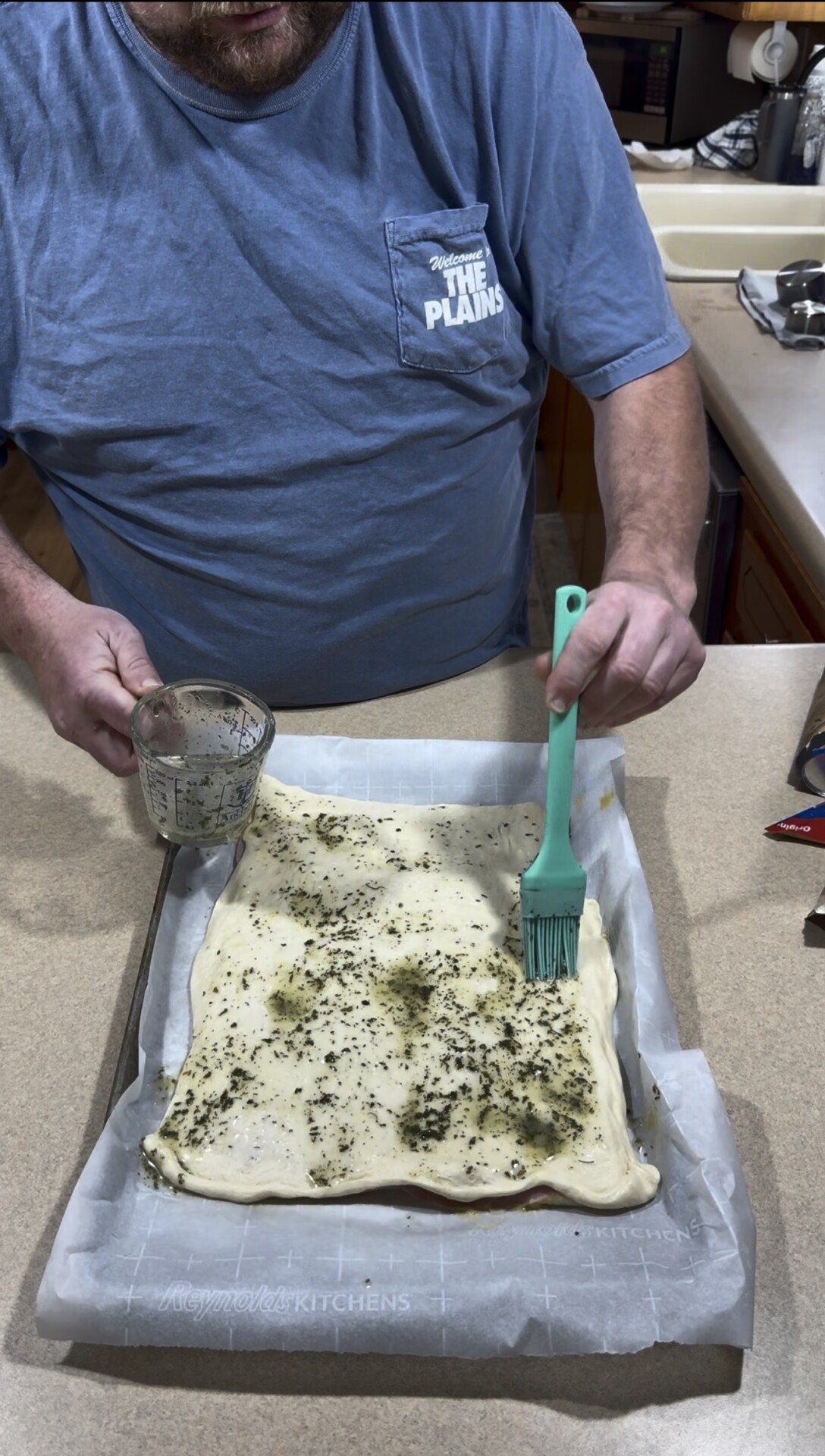 A person using a light green silicone brush to spread oil and herbs over a sheet of raw dough on a parchment-lined baking pan.