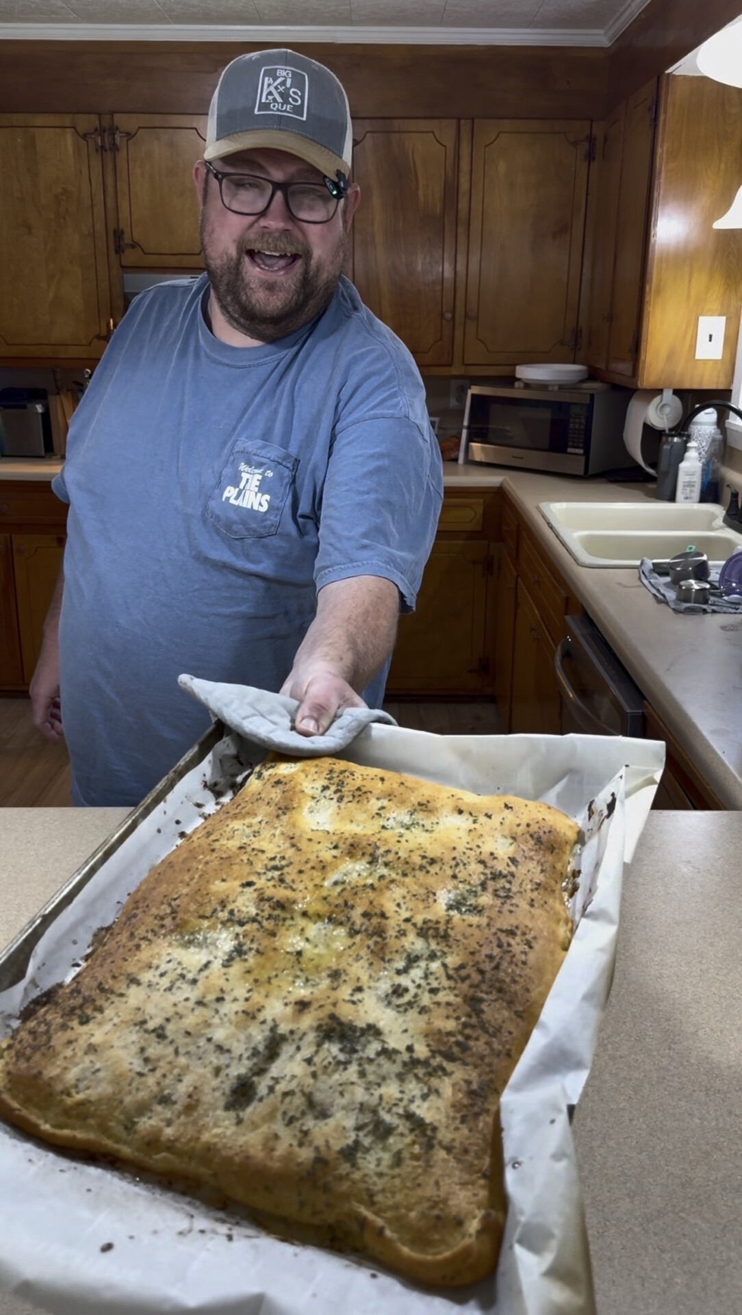 A man in a blue t-shirt and baseball cap holds a baking sheet with a large, golden-brown herb-topped flatbread.