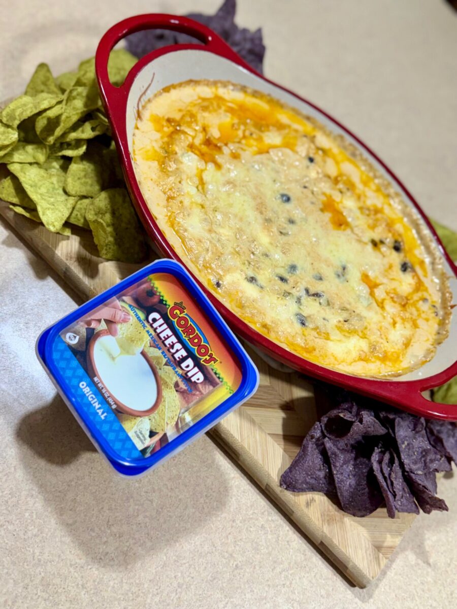 A red baking dish filled with melted cheese dip next to a container of Gordos cheese dip, surrounded by green and purple tortilla chips on a wooden board.