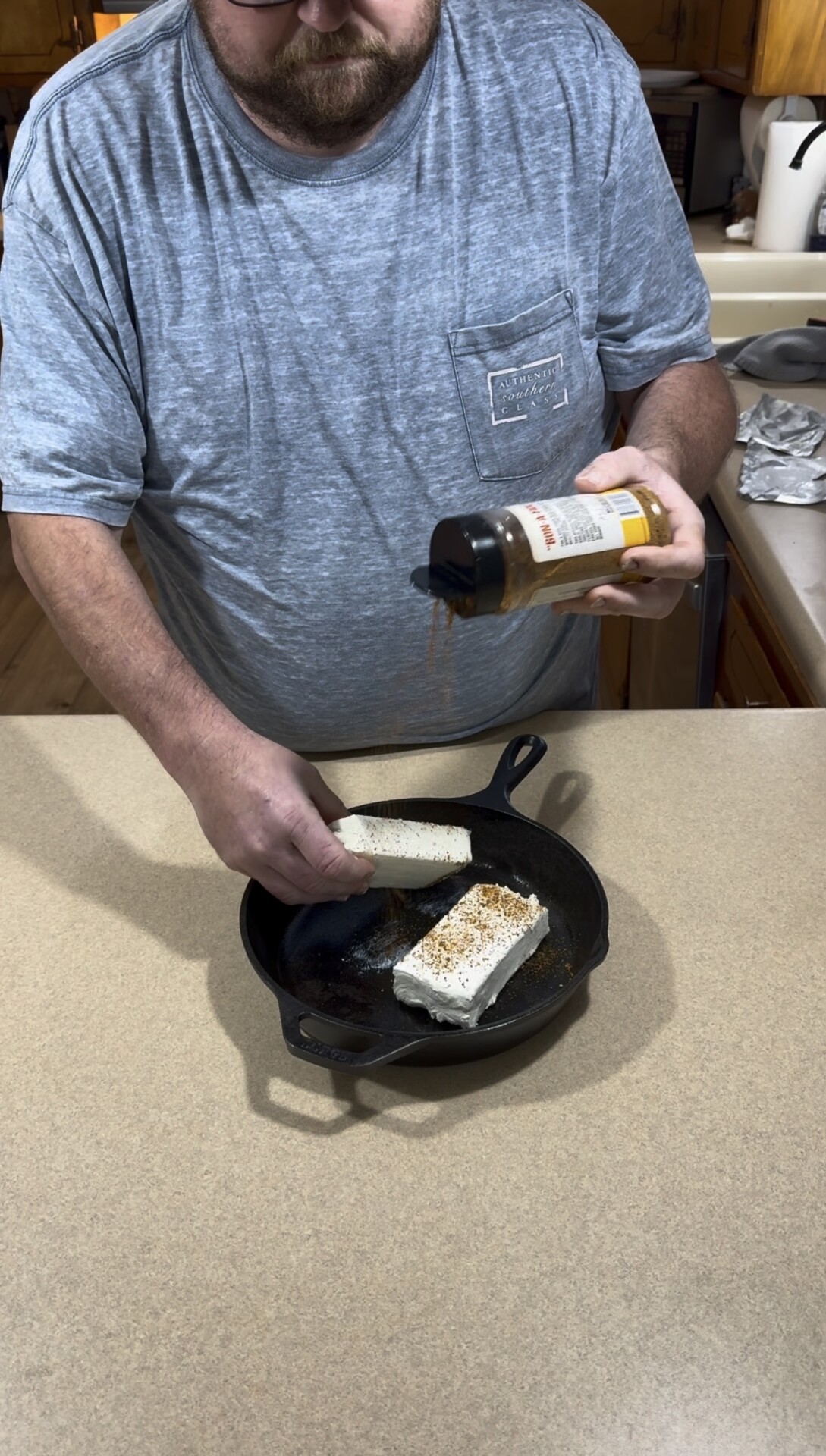 A person seasoning two blocks of cream cheese in a cast iron skillet.