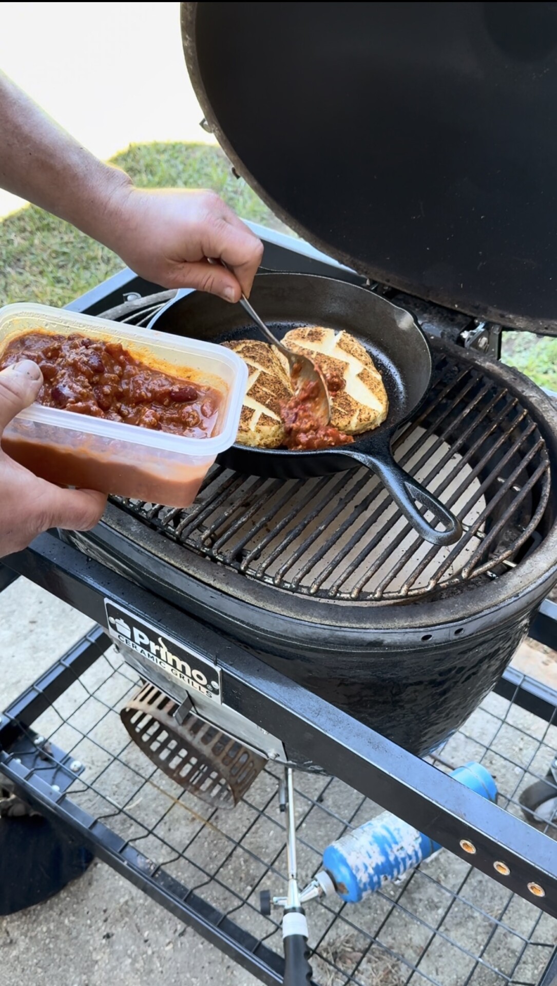A person uses a spoon to add chili from a container onto scored burger patties cooking in a cast iron skillet on a Primo ceramic grill.