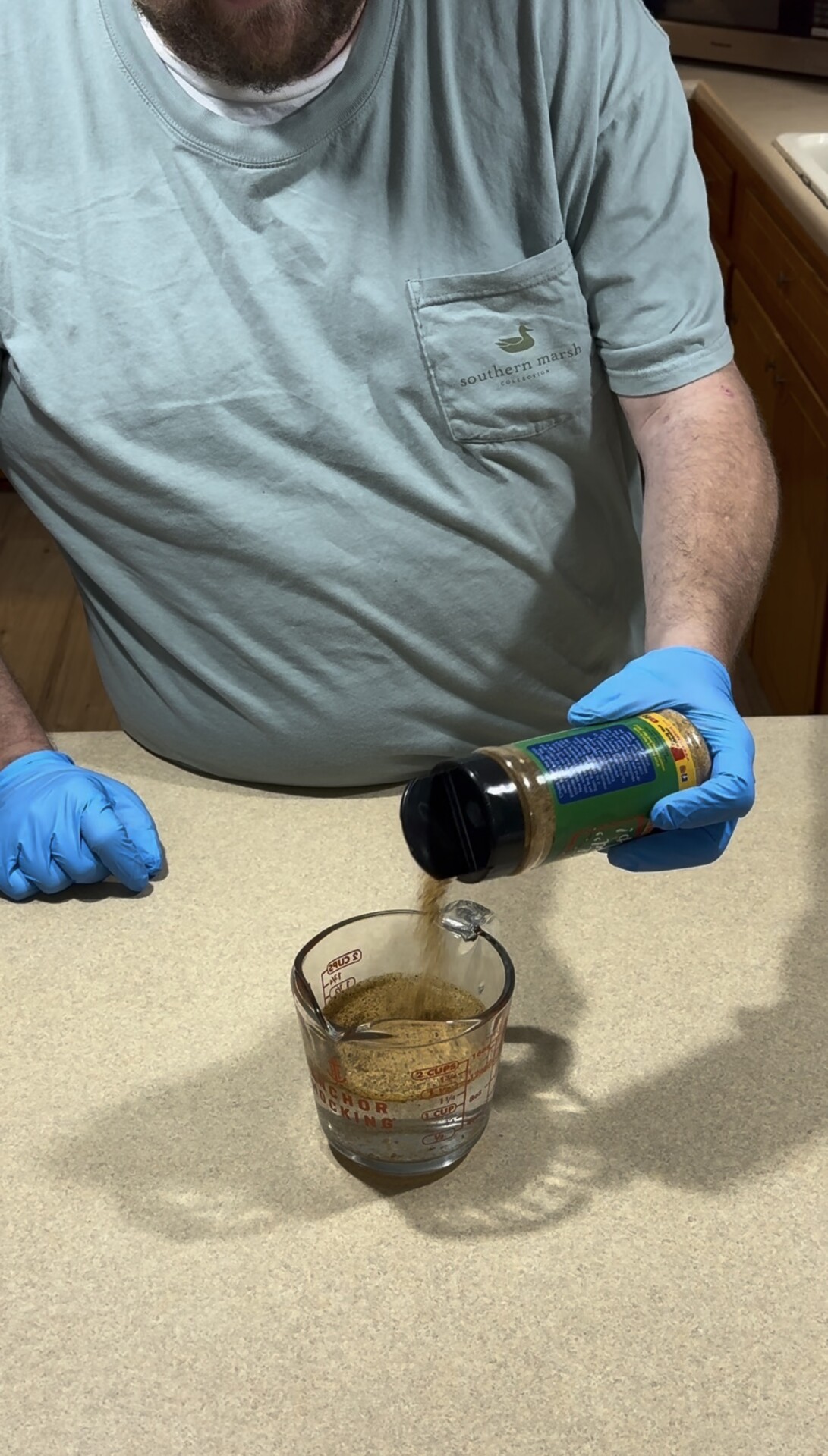 A person wearing blue gloves pours dry seasoning from a plastic shaker into a glass measuring cup.