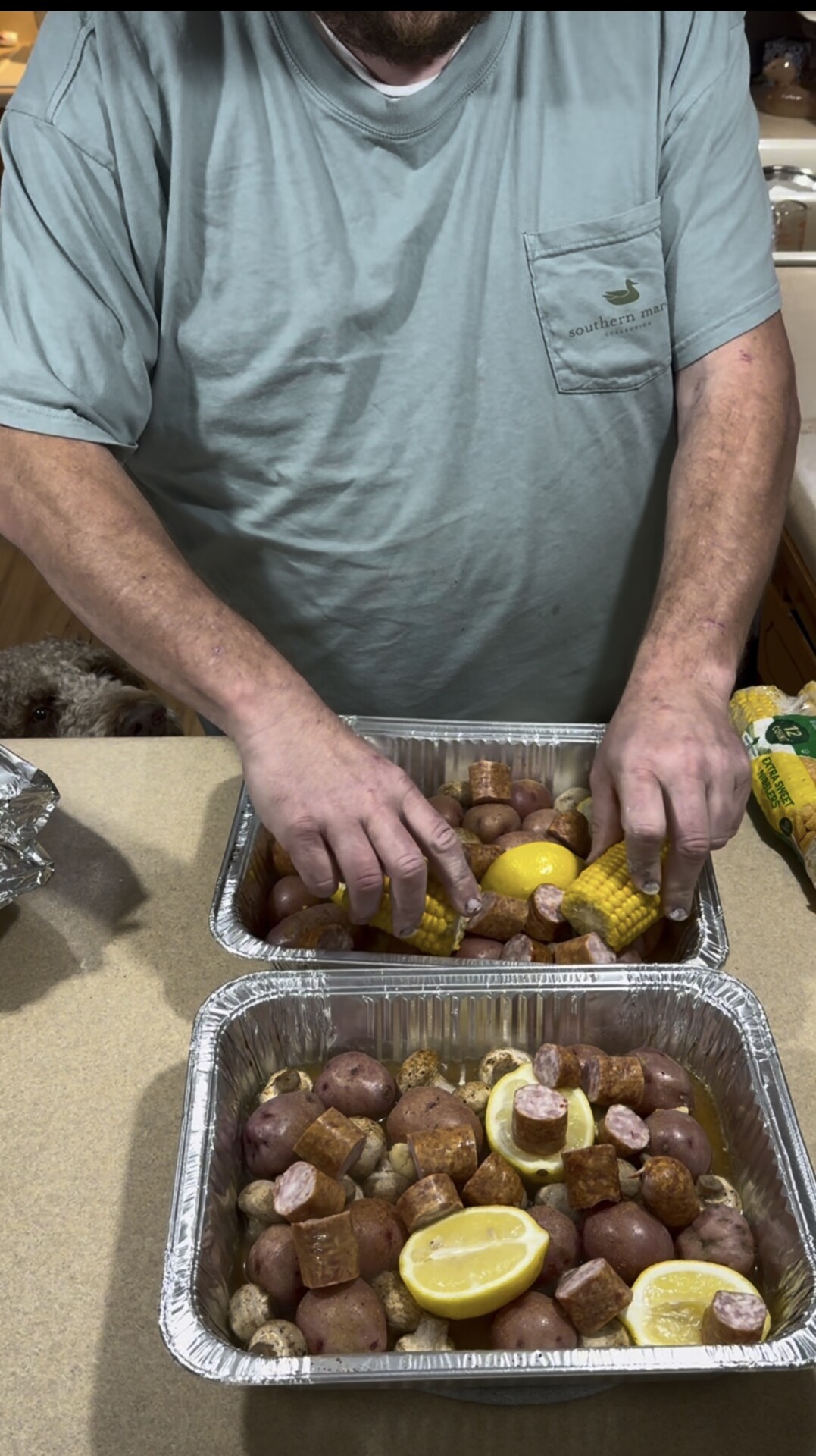 A person arranging corn, sausage, potatoes, and lemon halves in aluminum foil pans.
