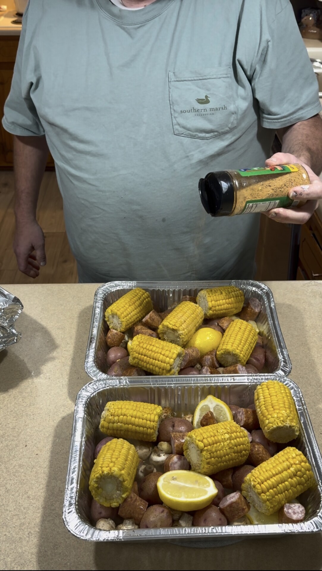 A person sprinkles seasoning over two foil pans filled with corn on the cob, sliced sausage, potatoes, and lemon halves.