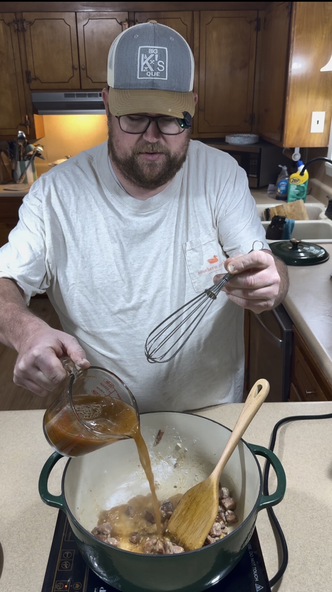 A man wearing a baseball cap and glasses pours broth from a measuring cup into a pot containing meat and a wooden spoon, holding a whisk in his other hand.