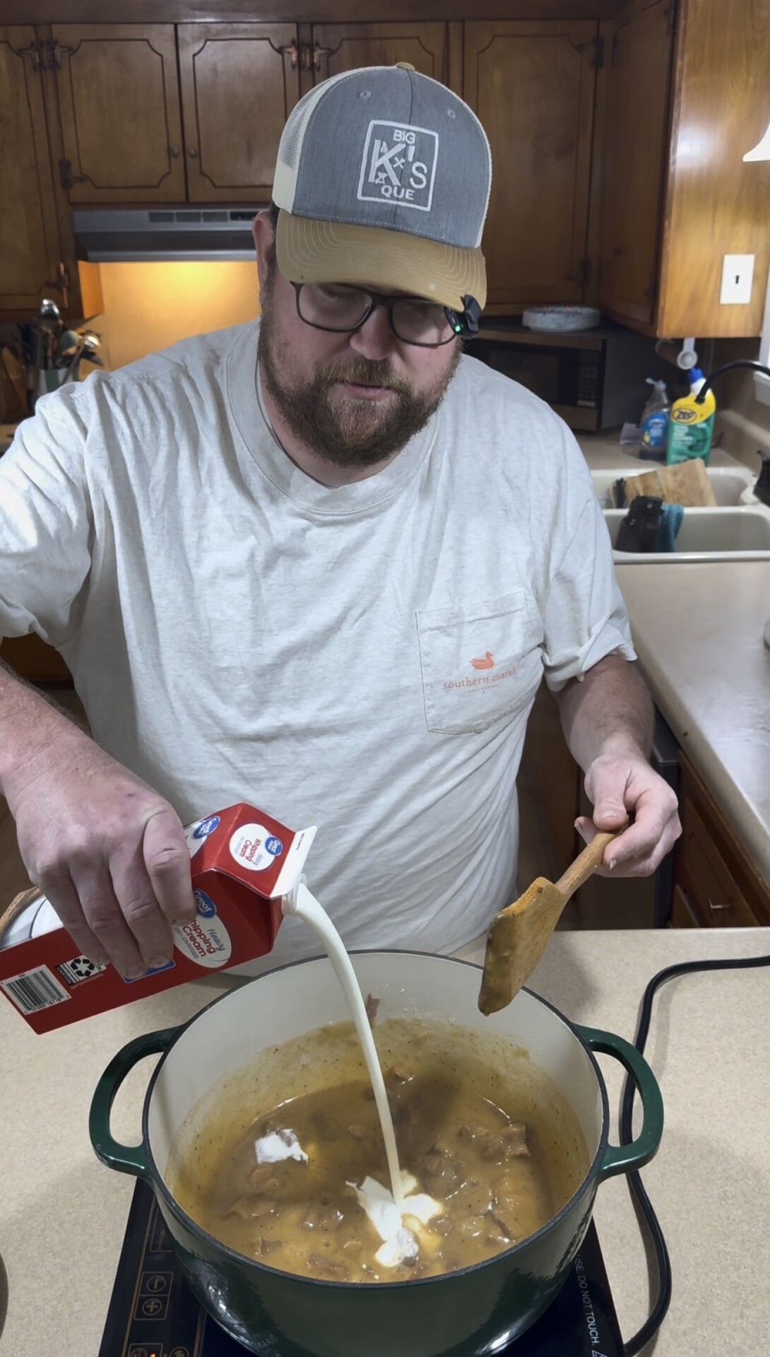 A man in a baseball cap pouring heavy whipping cream into a pot of stew on a kitchen counter.