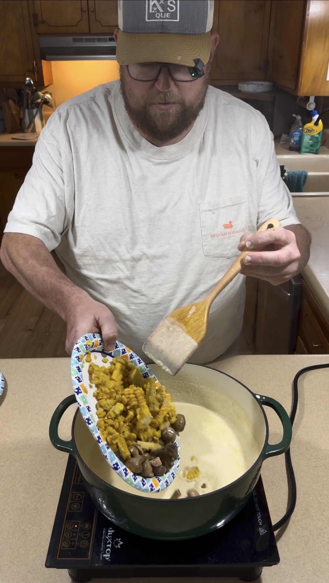 A person pouring corn and mushrooms from a paper plate into a pot of cream-based soup on an induction cooktop.