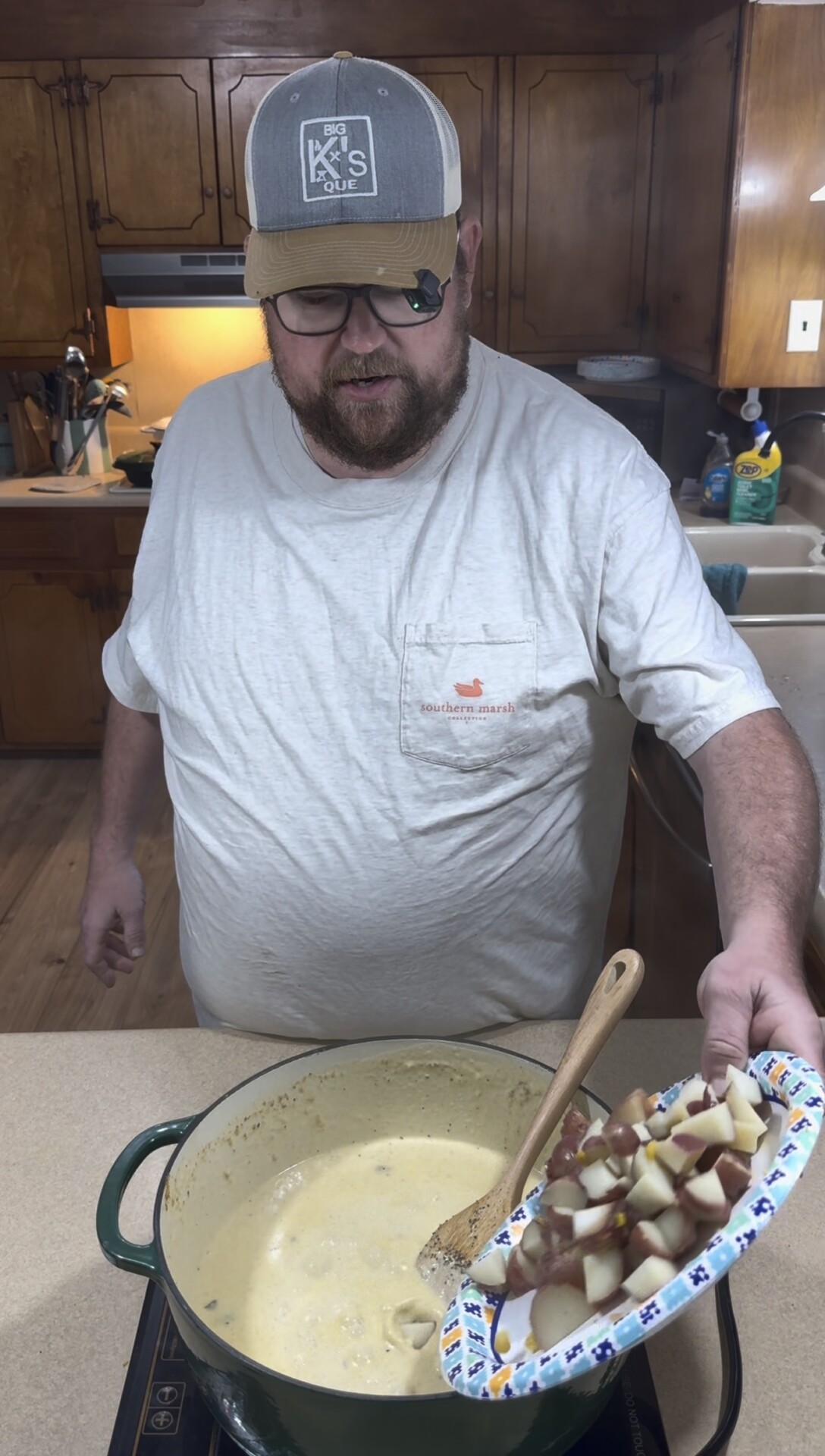 A man wearing glasses and a baseball cap adds chopped potatoes from a paper plate into a pot of cream-based soup on a stove.