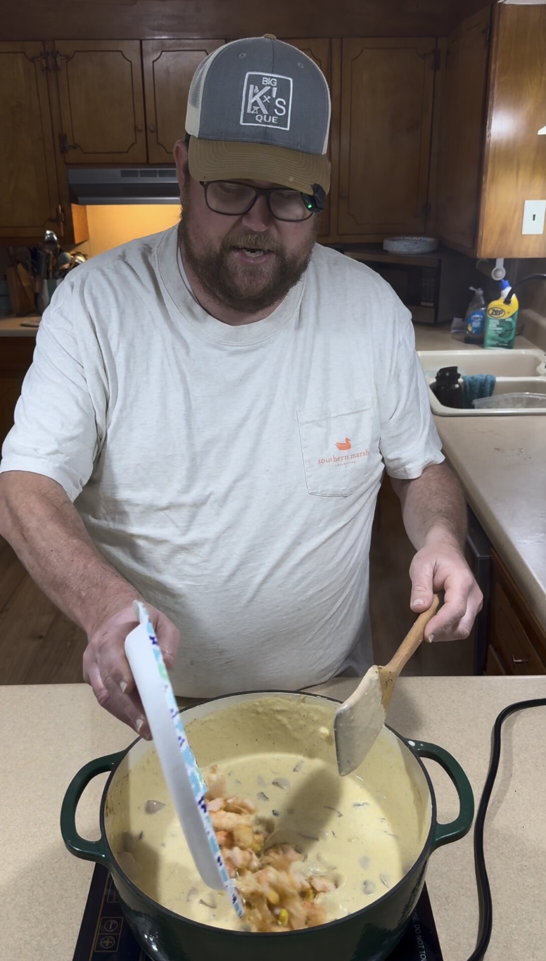 A man in a baseball cap and white t-shirt uses a wooden spoon to stir a pot of creamy sauce as he adds a bowl of shrimp to the mixture.
