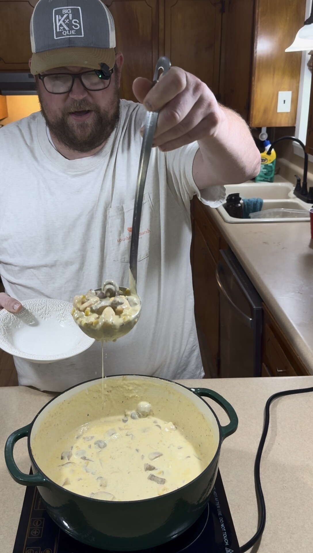 A man in a baseball cap holding a ladle of creamy corn and mushroom soup over a large pot.