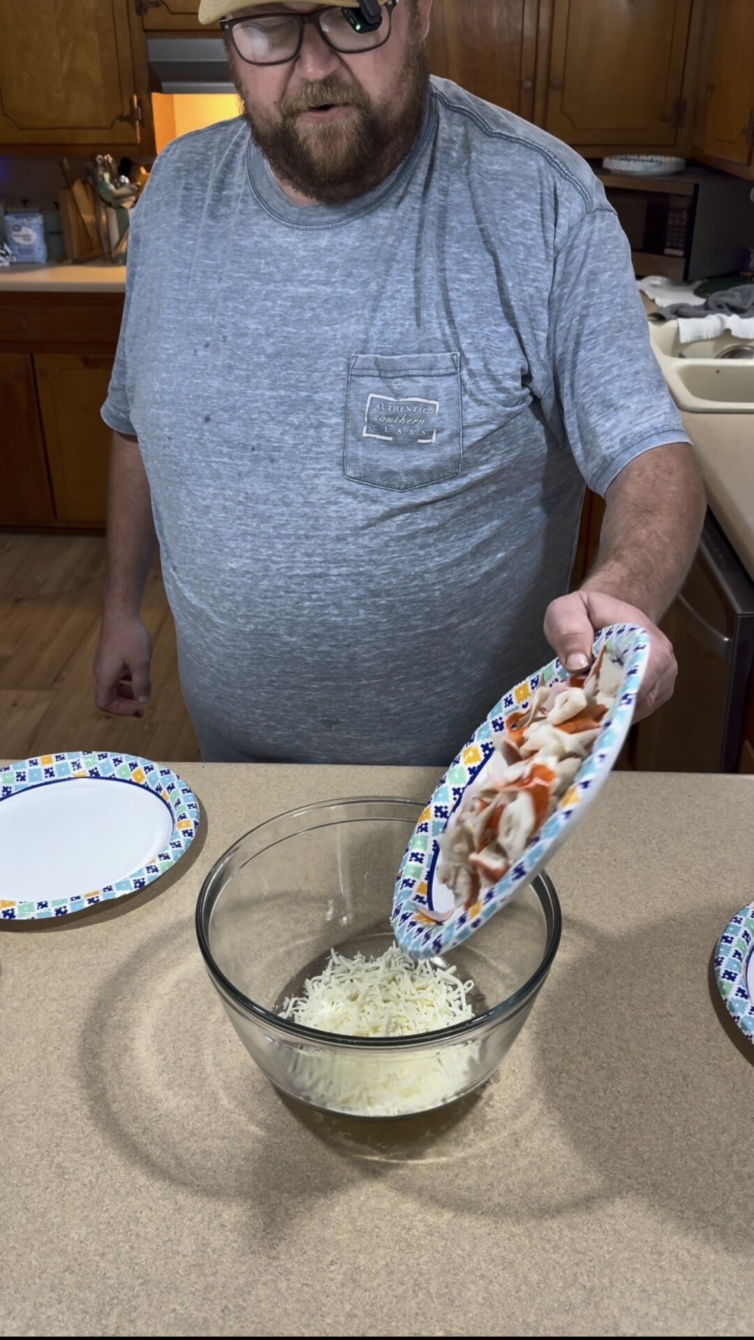 A man holding a paper plate pours sliced pepperoni and meat into a glass bowl containing shredded cheese.