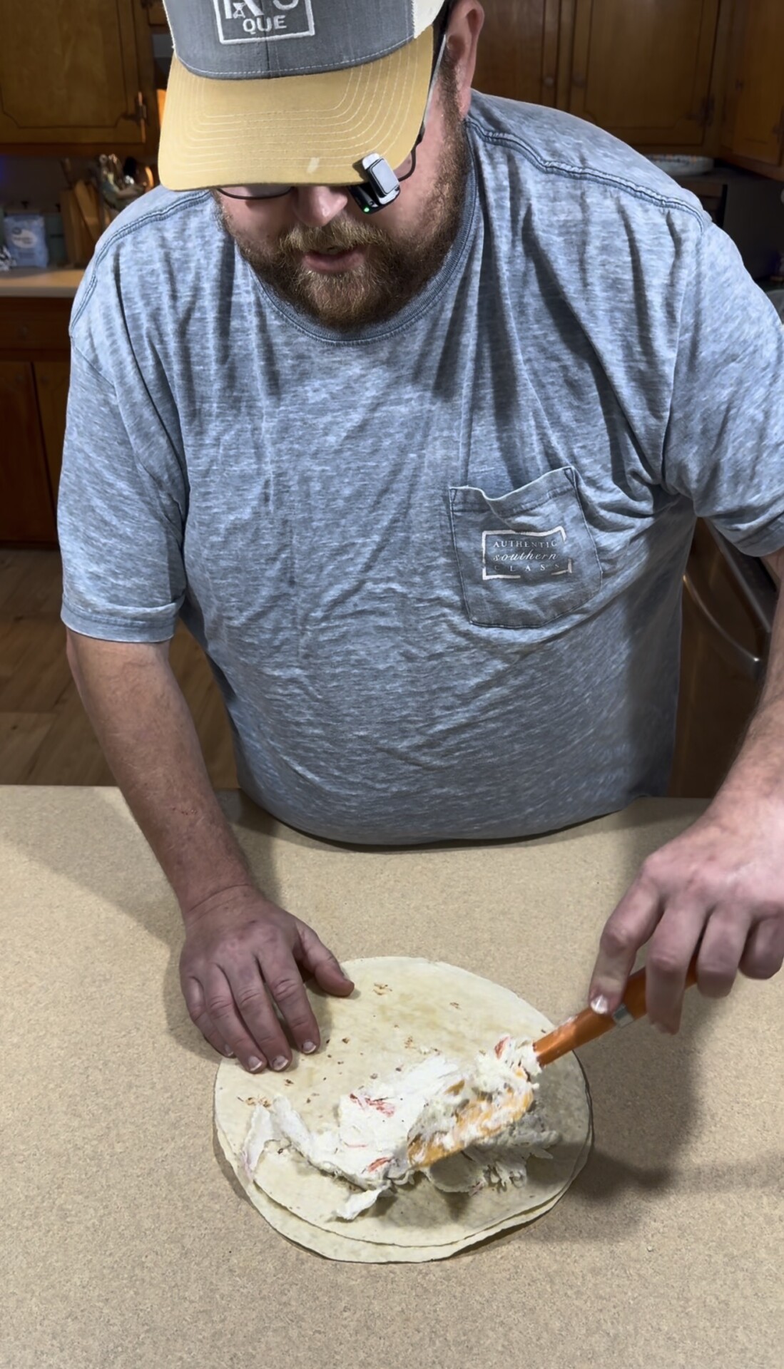 A man wearing a baseball cap spreads a creamy dip onto a flour tortilla with a spatula.