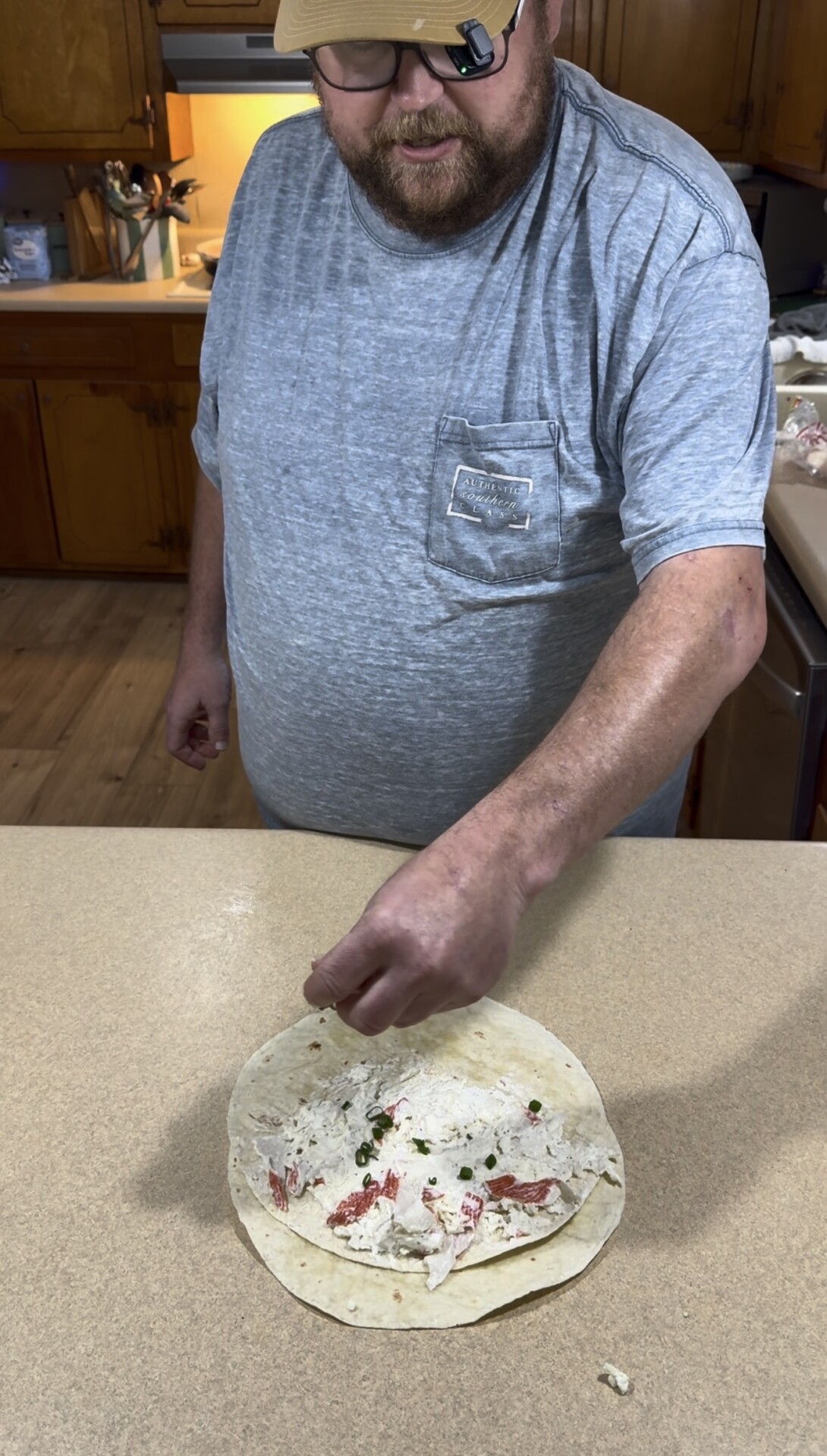 A man with a beard and baseball cap sprinkles green onions over a tortilla topped with a crab and cream cheese mixture.