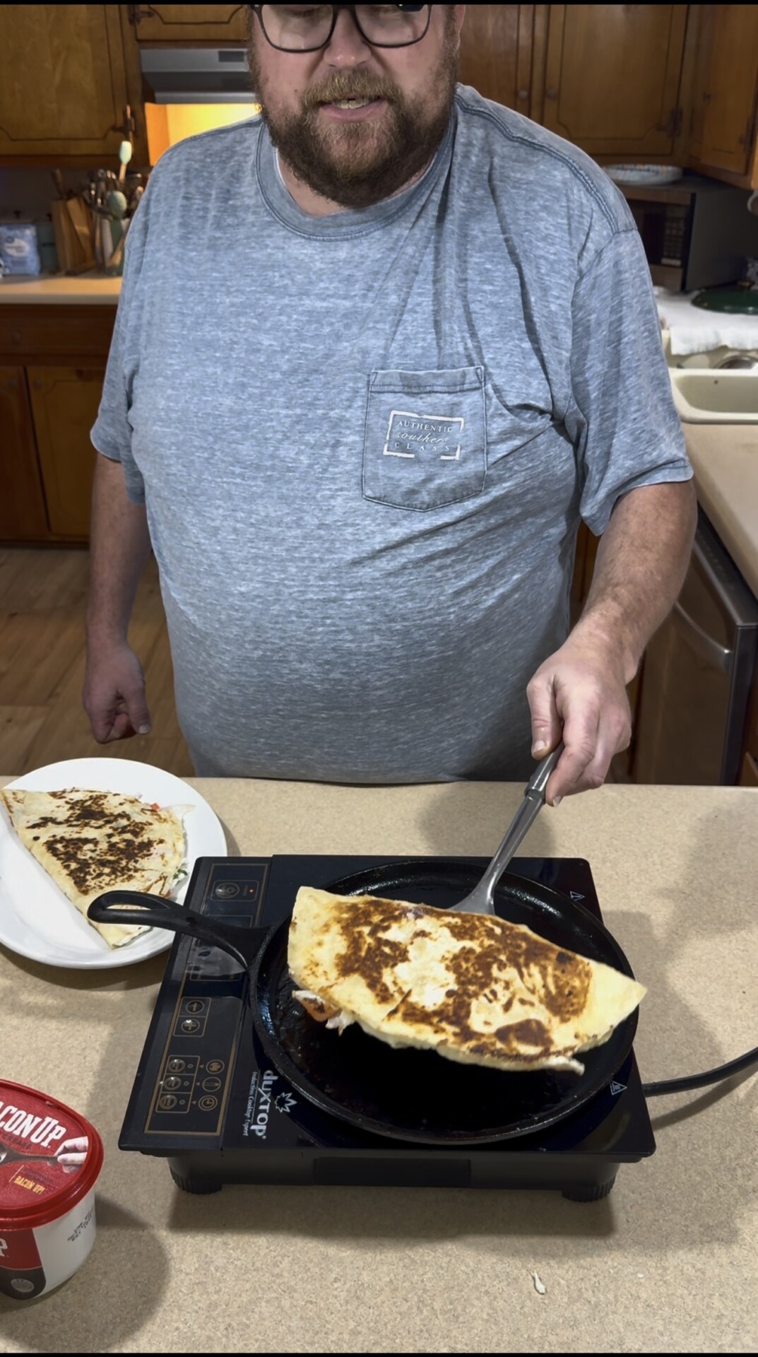 A man in a grey t-shirt flips a toasted quesadilla in a cast iron skillet on a portable induction cooktop.