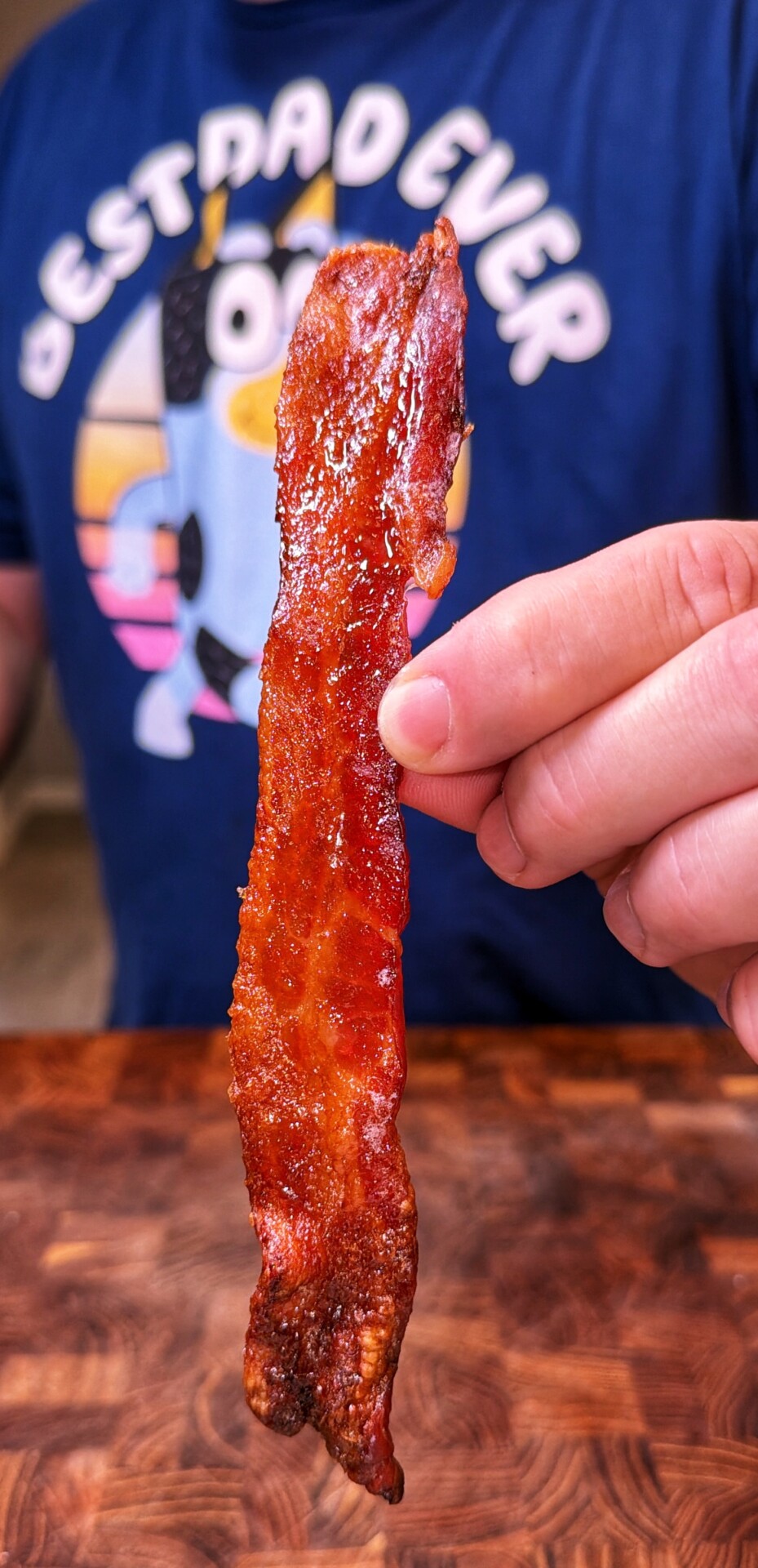 A person holds a crispy strip of bacon in front of their chest, wearing a blue Best Dad Ever t-shirt with a cartoon character on it.