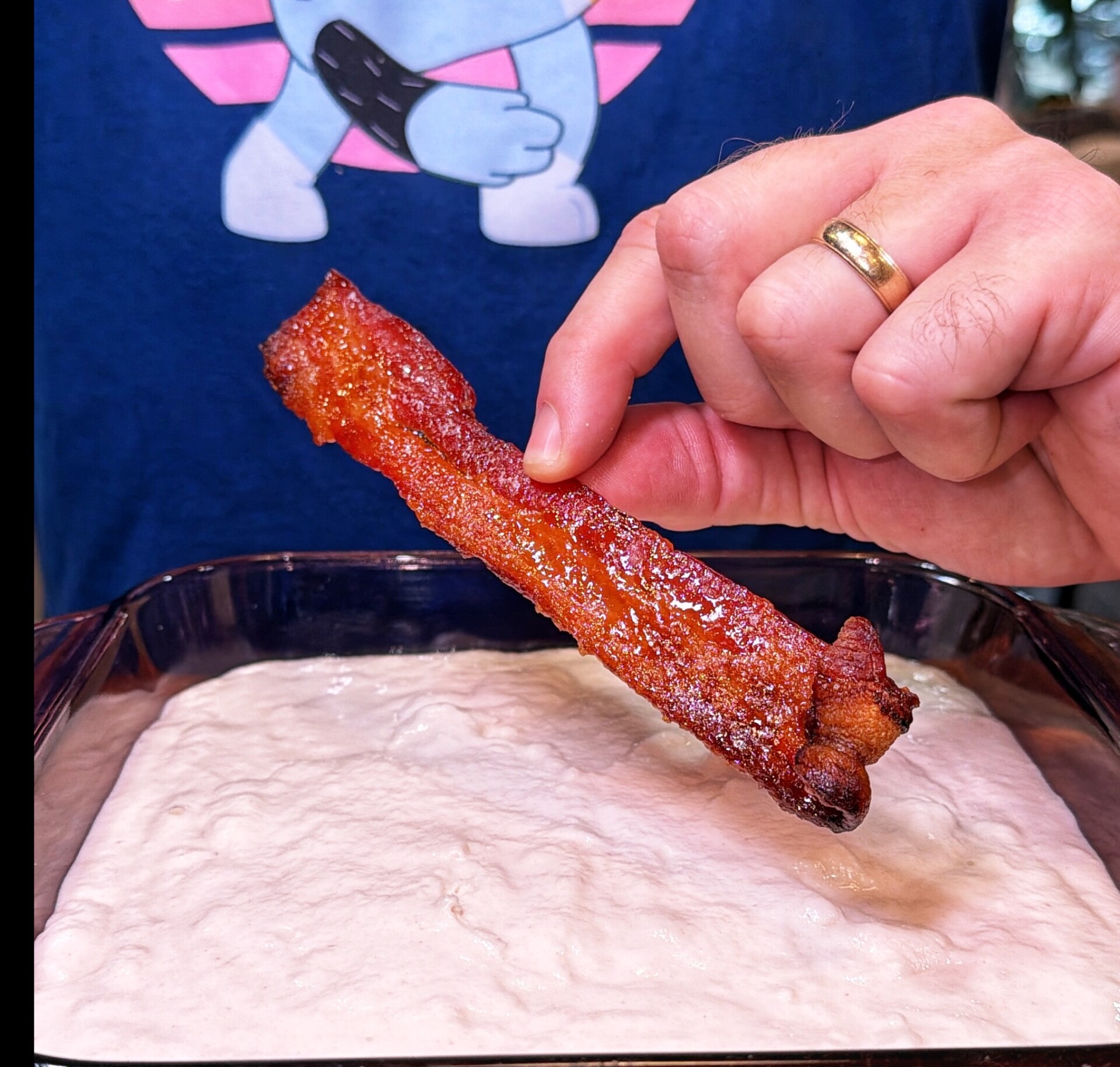 A hand holding a crispy piece of bacon over a dish of white dip, with a person in a blue shirt featuring a cartoon graphic in the background.