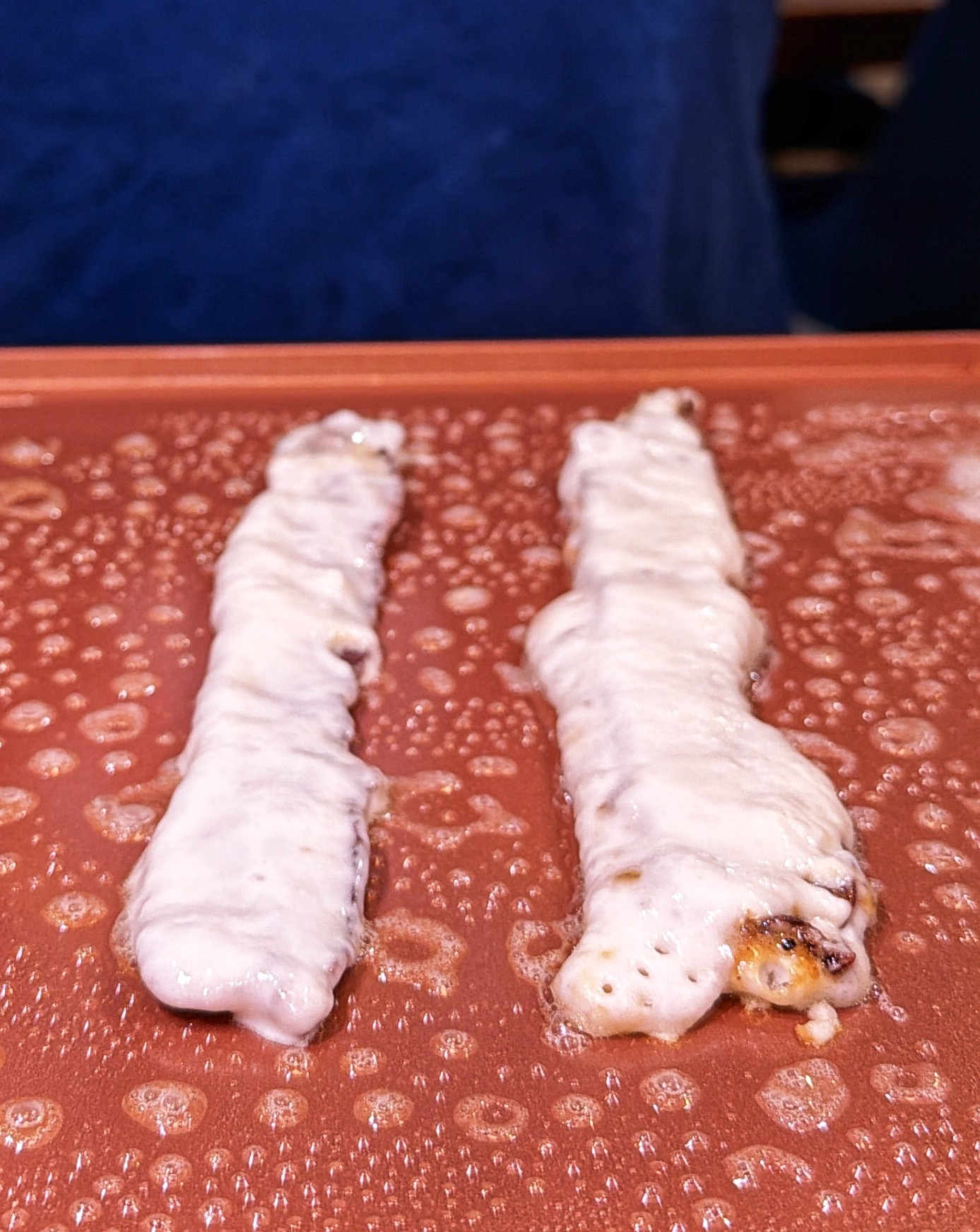 Two skewers covered in melted cheese are lying on a sizzling brown grilling surface, with bubbles and steam visible around them.