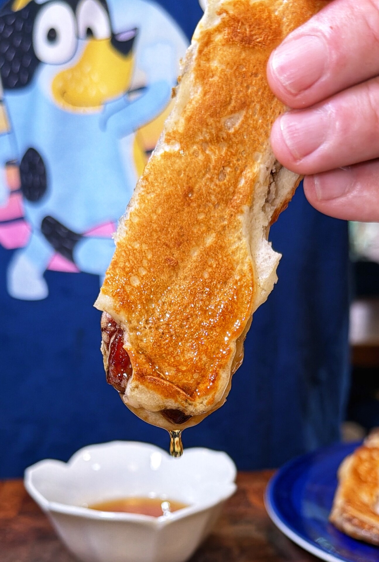 A hand holds a syrup-dripping pancake stick above a small bowl of syrup. A blue shirt with a cartoon dog character is visible in the background.