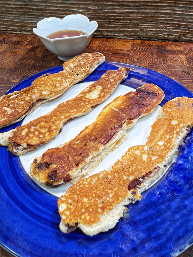 Long, rectangular pancakes on a blue plate, served with a small bowl of syrup in the background on a wooden surface.