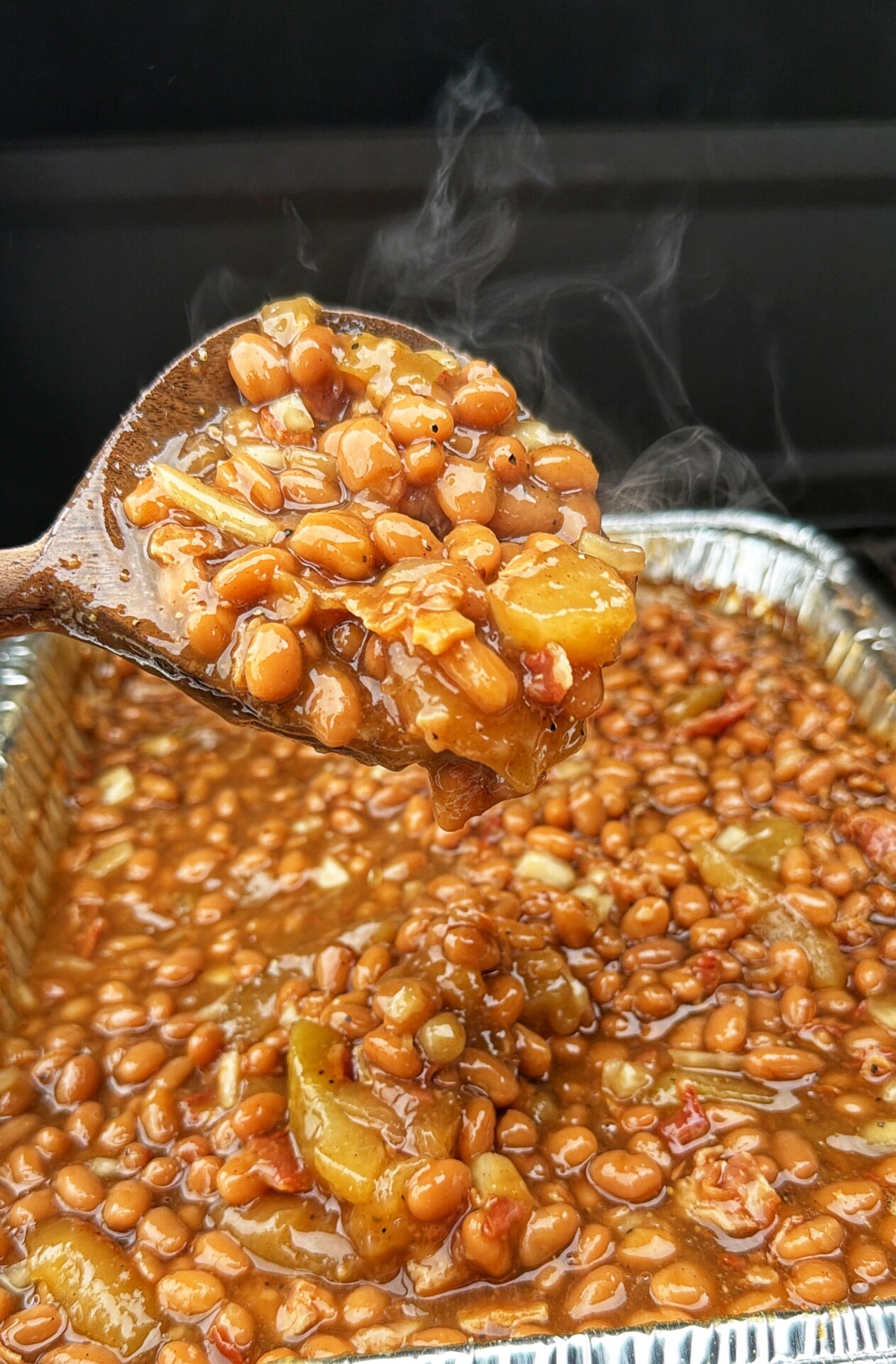 A wooden spoon lifts steaming baked beans with chunks of peppers and onions from a foil tray filled with baked beans.