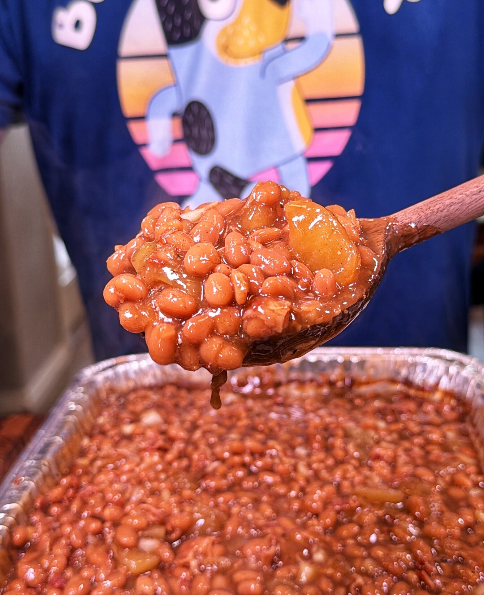 A wooden spoon lifts baked beans from a foil tray. The background shows a person wearing a blue shirt with a cartoon graphic.