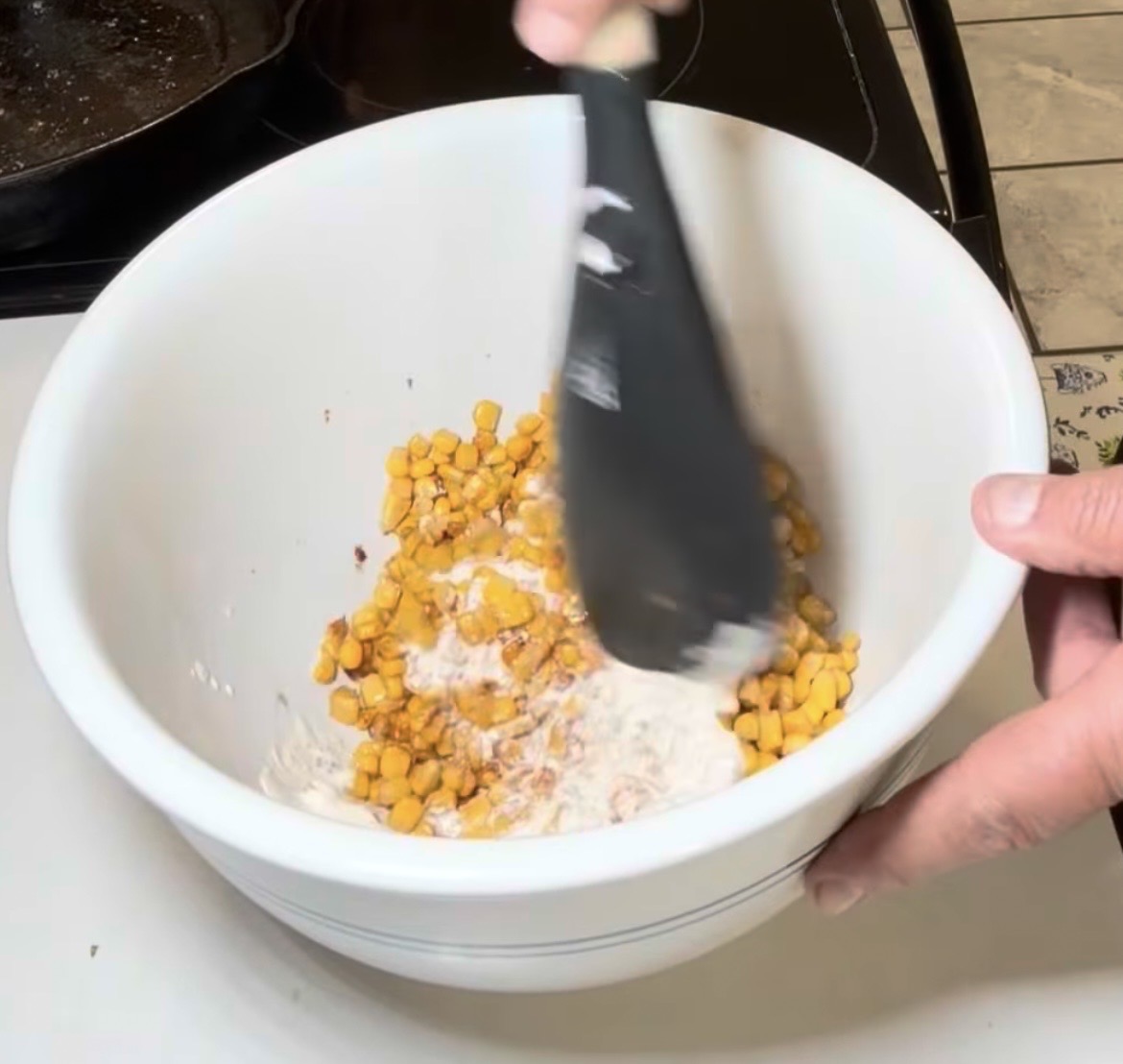 A hand uses a spatula to stir corn kernels into a white creamy sauce in a white bowl.