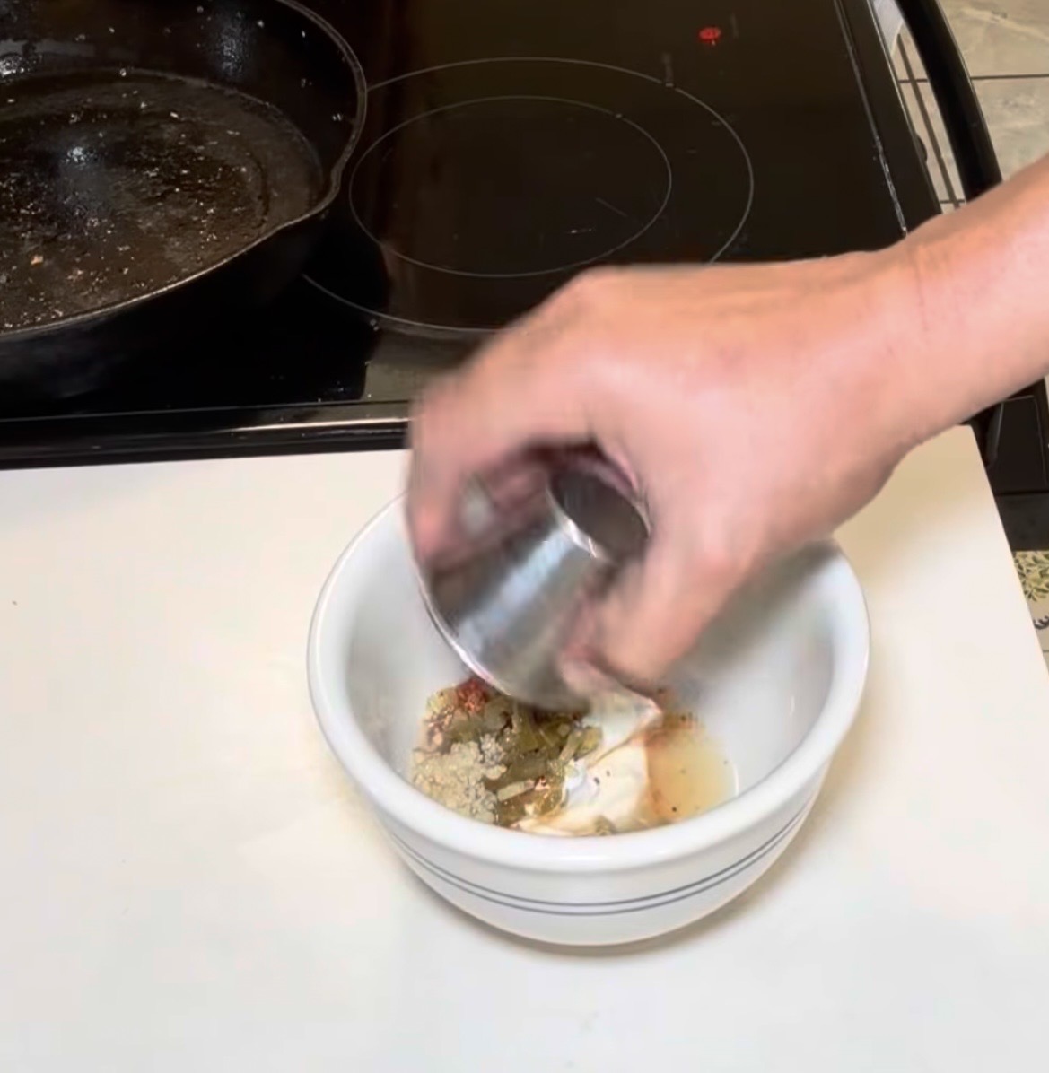 A hand pours liquid from a metal jigger into a white bowl containing white sauce, spices, and chopped green herbs.