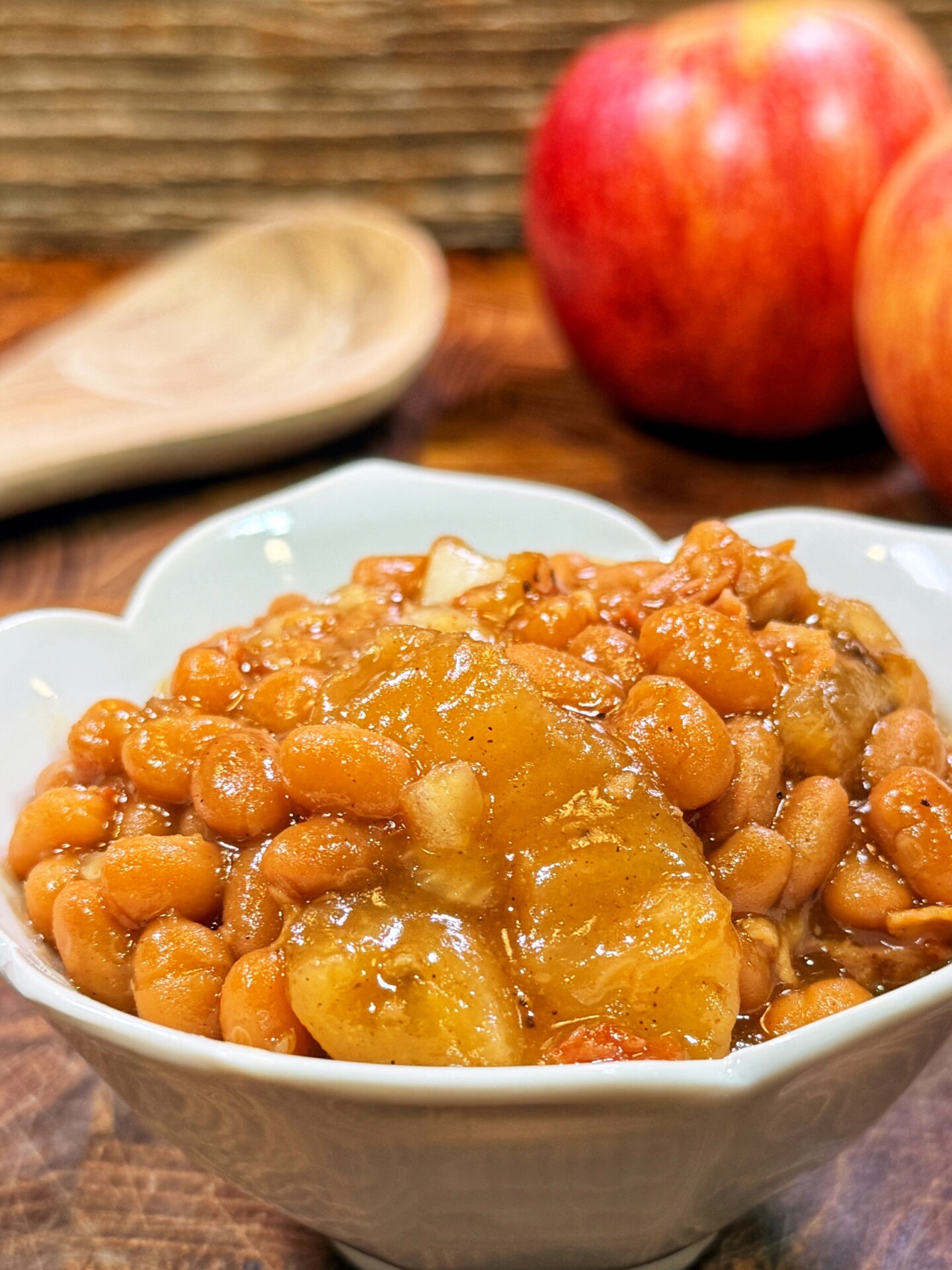 A white heart-shaped bowl filled with baked beans and chunks of apple, placed on a wooden surface with two apples and a wooden spoon in the background.