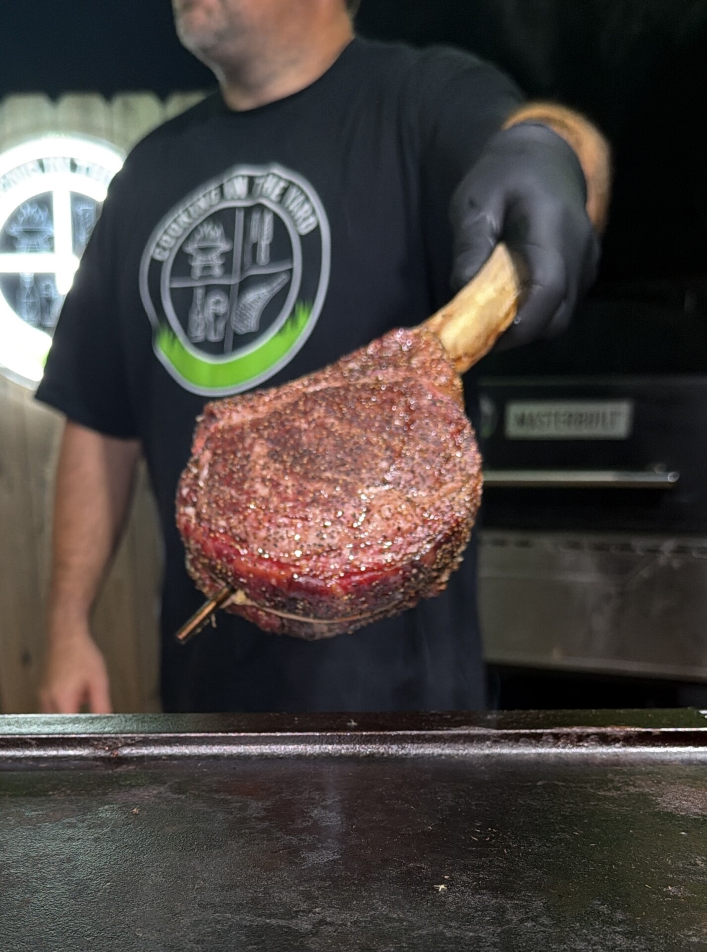 A person wearing black gloves holds a large, seasoned tomahawk steak over a grill.