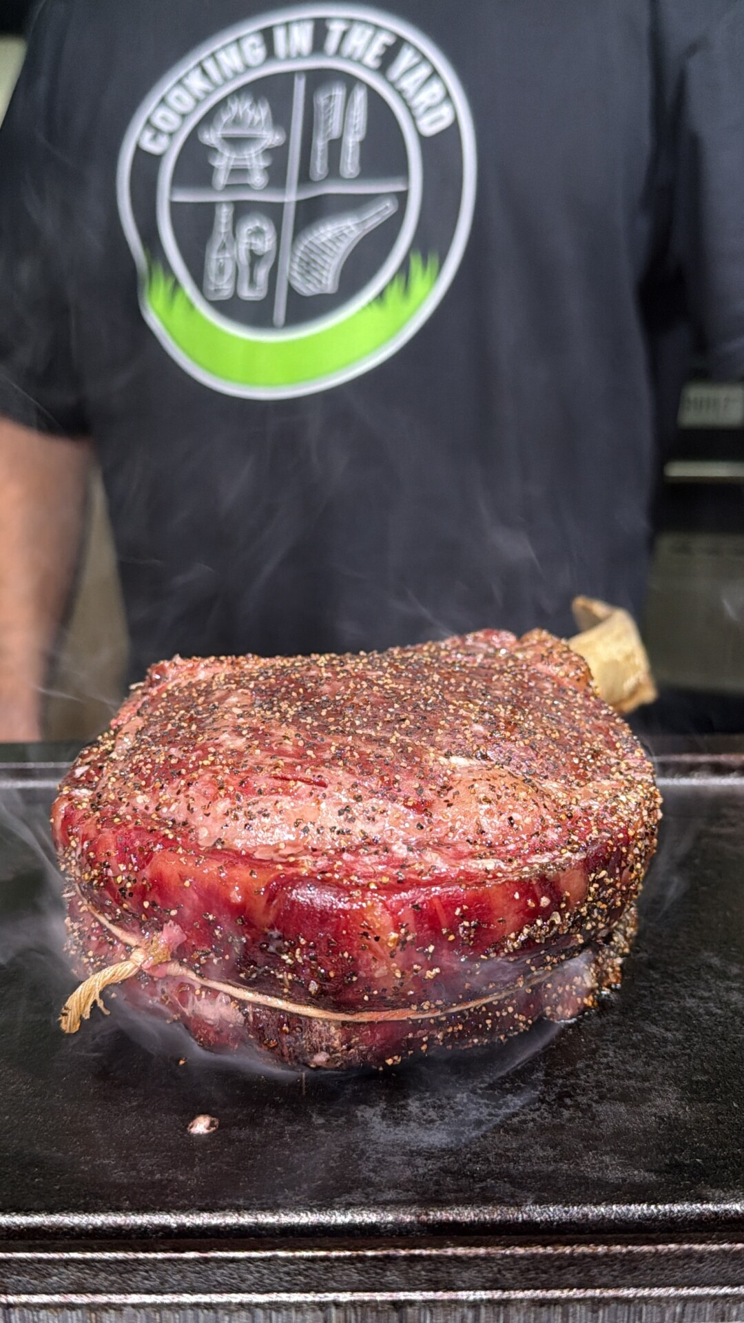 A person wearing a Cooking in the Yard shirt stands behind a large, pepper-crusted, bone-in steak searing on a hot grill.