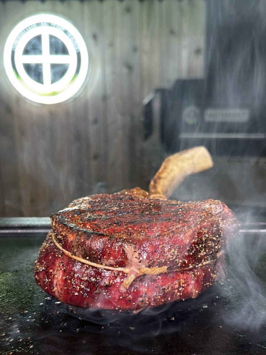 A seasoned tomahawk steak searing on a hot flat-top grill with smoke rising around it.