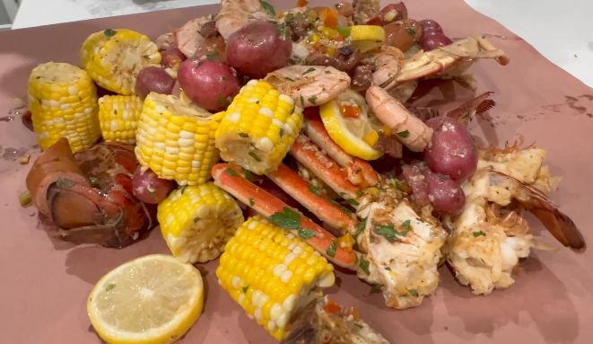 A seafood boil spread on butcher paper featuring crab legs, lobster, shrimp, corn on the cob, red potatoes, and lemon slices.