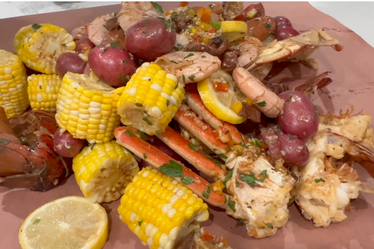 A seafood boil featuring snow crab legs, shrimp, corn on the cob, and red potatoes, garnished with lemon slices and herbs.